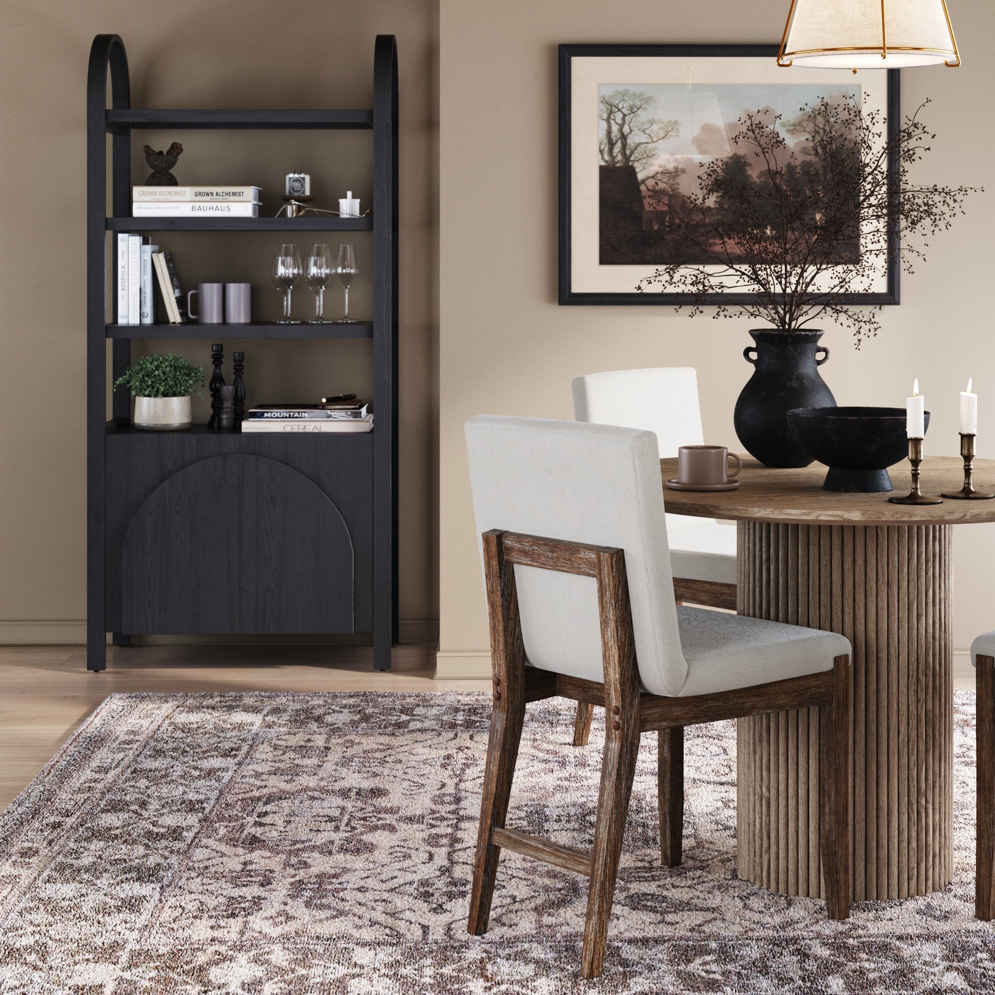 A modern dining room with a round wooden table, cushioned chairs, a patterned rug, framed art on a light brown wall, candles, a black vase with branches, and the Nathan James Mid-Century Arched Door Bookshelf with Doors Black displaying decor and books.