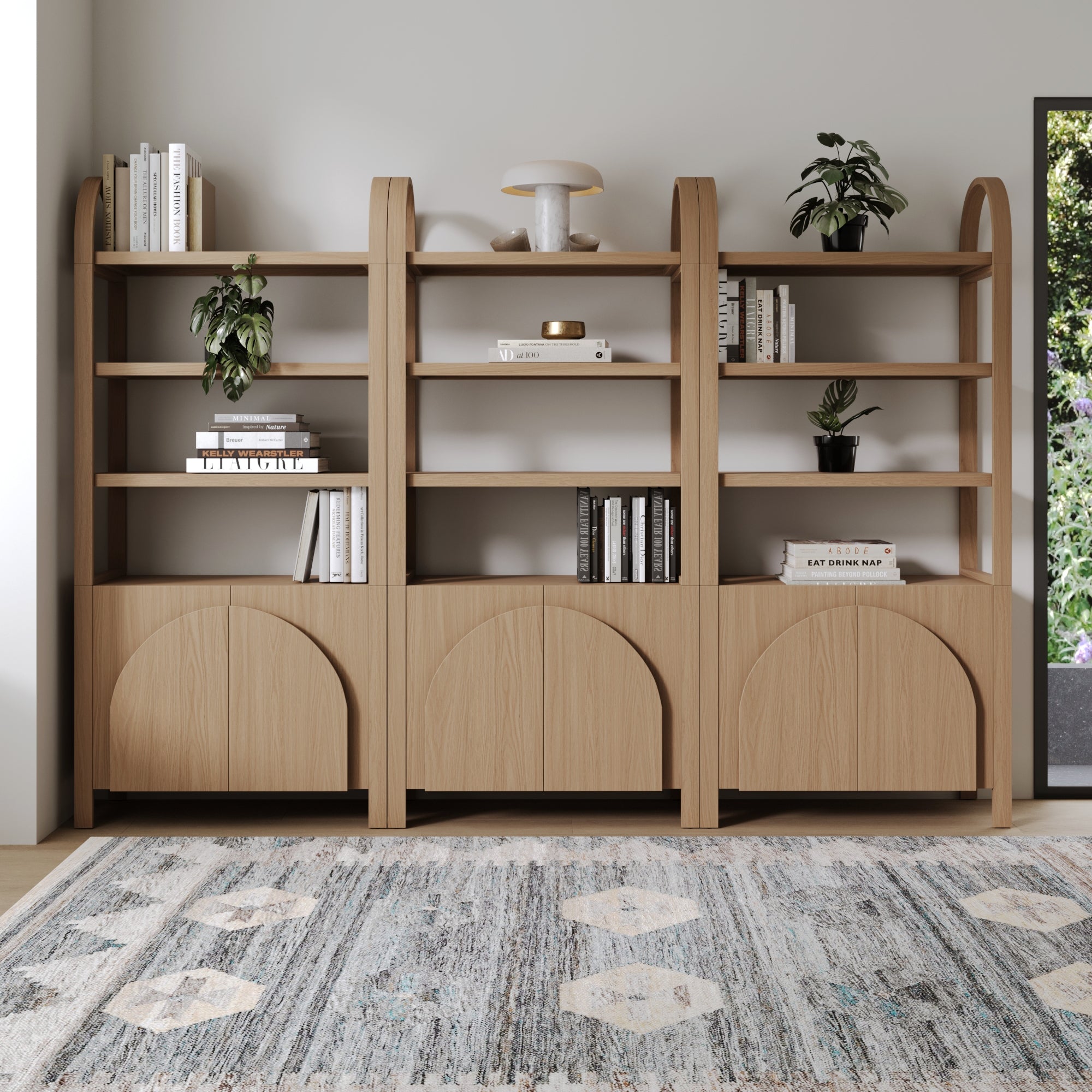 Three light wood bookshelves with minimalist arched tops stand against a white wall. They hold books, potted plants, and decor items. A patterned rug covers the floor, and sunlight shines from the right side.
