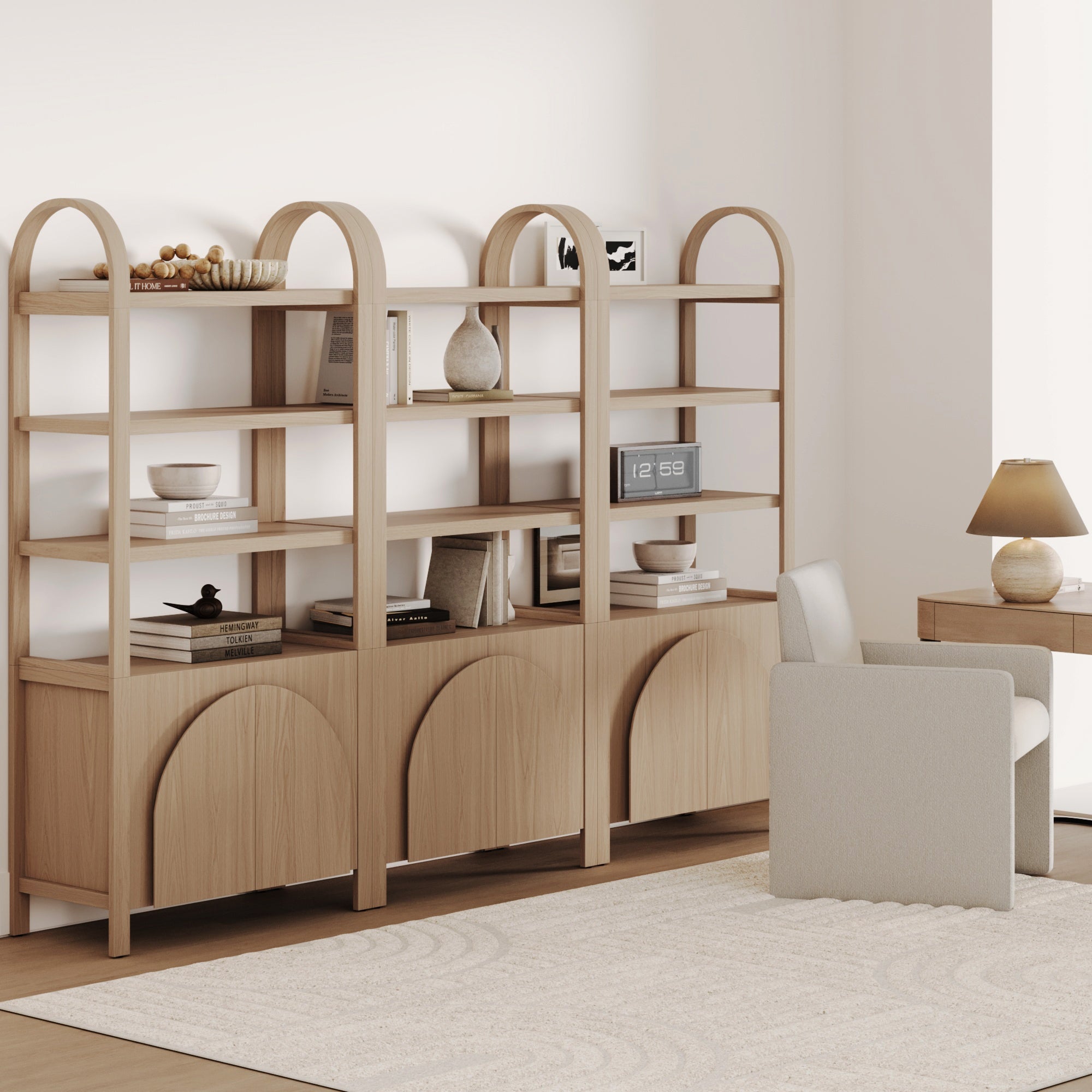 A modern home office features the Nathan James Mid-Century Arched Door Bookshelf with Doors Raw Oak (Set of 3), neutral decor, a desk with a lamp, and a plush cream chair on a soft rug for an airy, minimal look in natural tones.