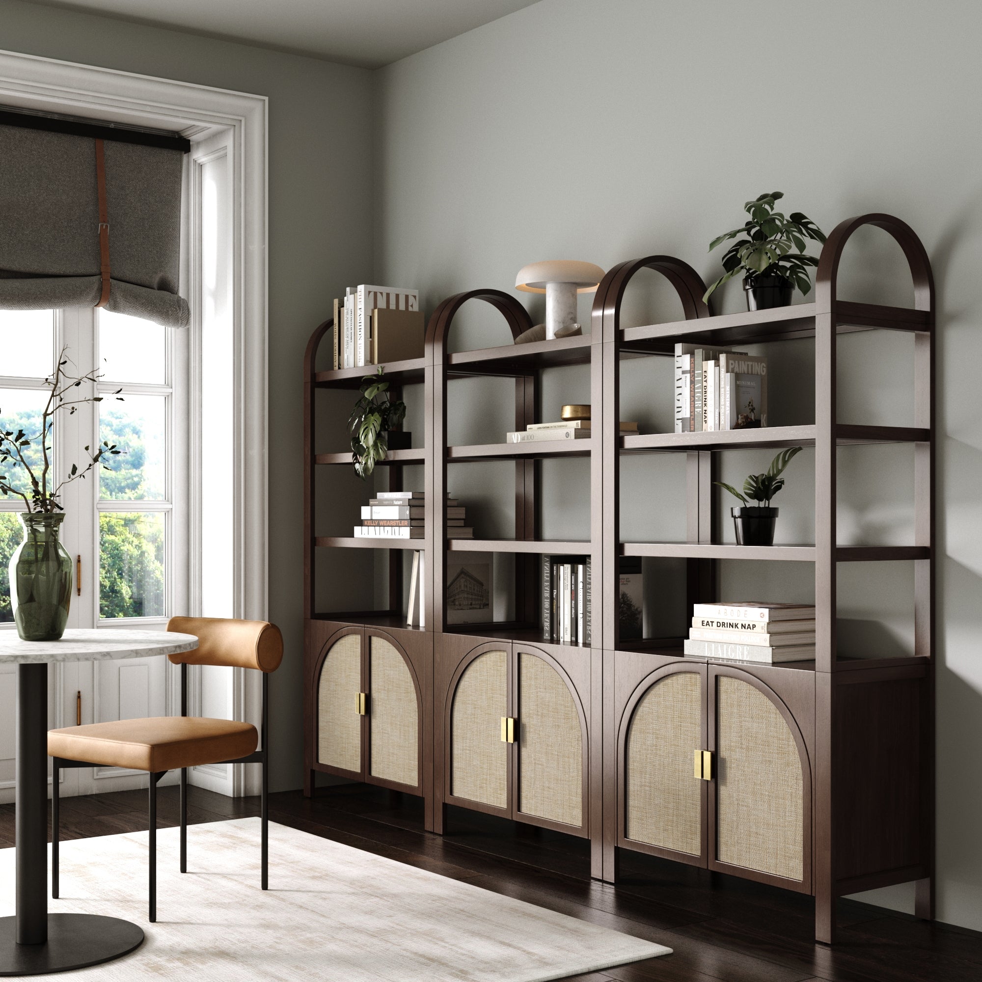 A modern home office featuring three Nathan James Arched Door Rattan Bookshelves with doors in Dark Acacia, styled with plants, books, decor, brown storage cabinets below, a tan chair, a round table, and a window with a gray Roman shade.