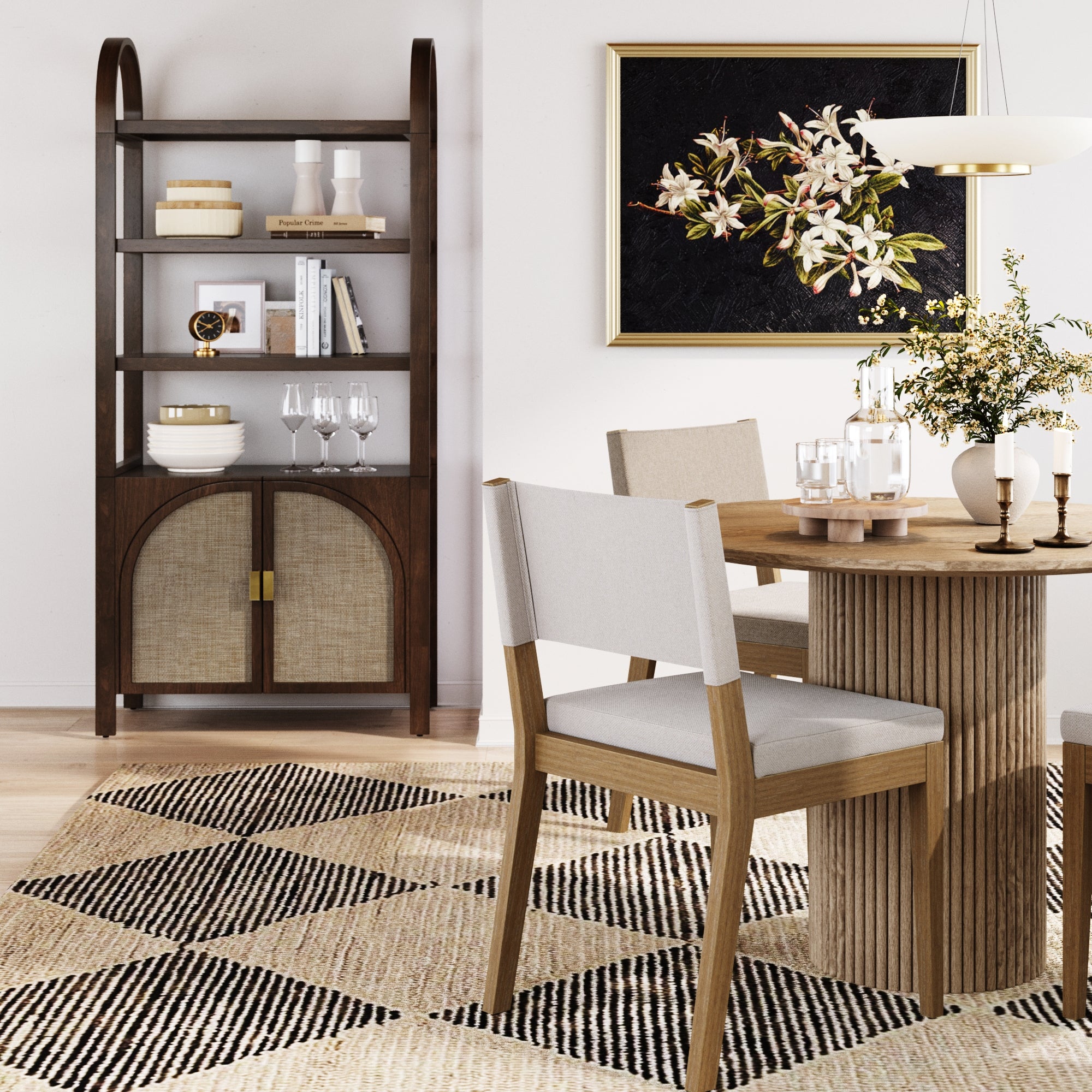 A modern dining room features a solid wood table, light upholstered chairs, a patterned rug, and the Nathan James Arched Door Rattan Bookshelf with Doors in Dark Acacia adorned with decor. A framed floral artwork and a small vase with greenery complete the look.