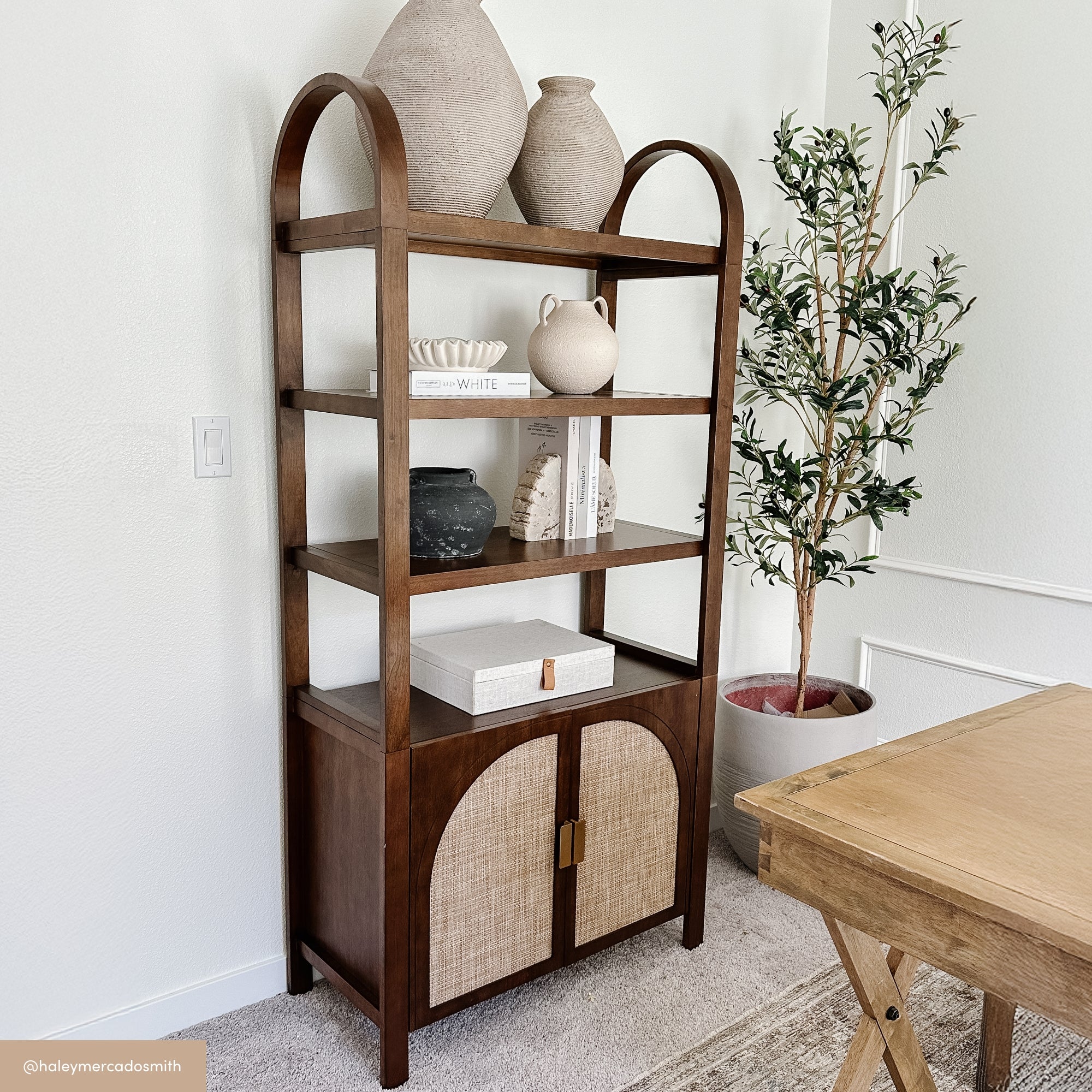 A tall wooden bookshelf with arched sides displays decorative vases, books, and pottery. It stands against a light wall beside a potted plant and a wooden table in a bright, stylish room.
