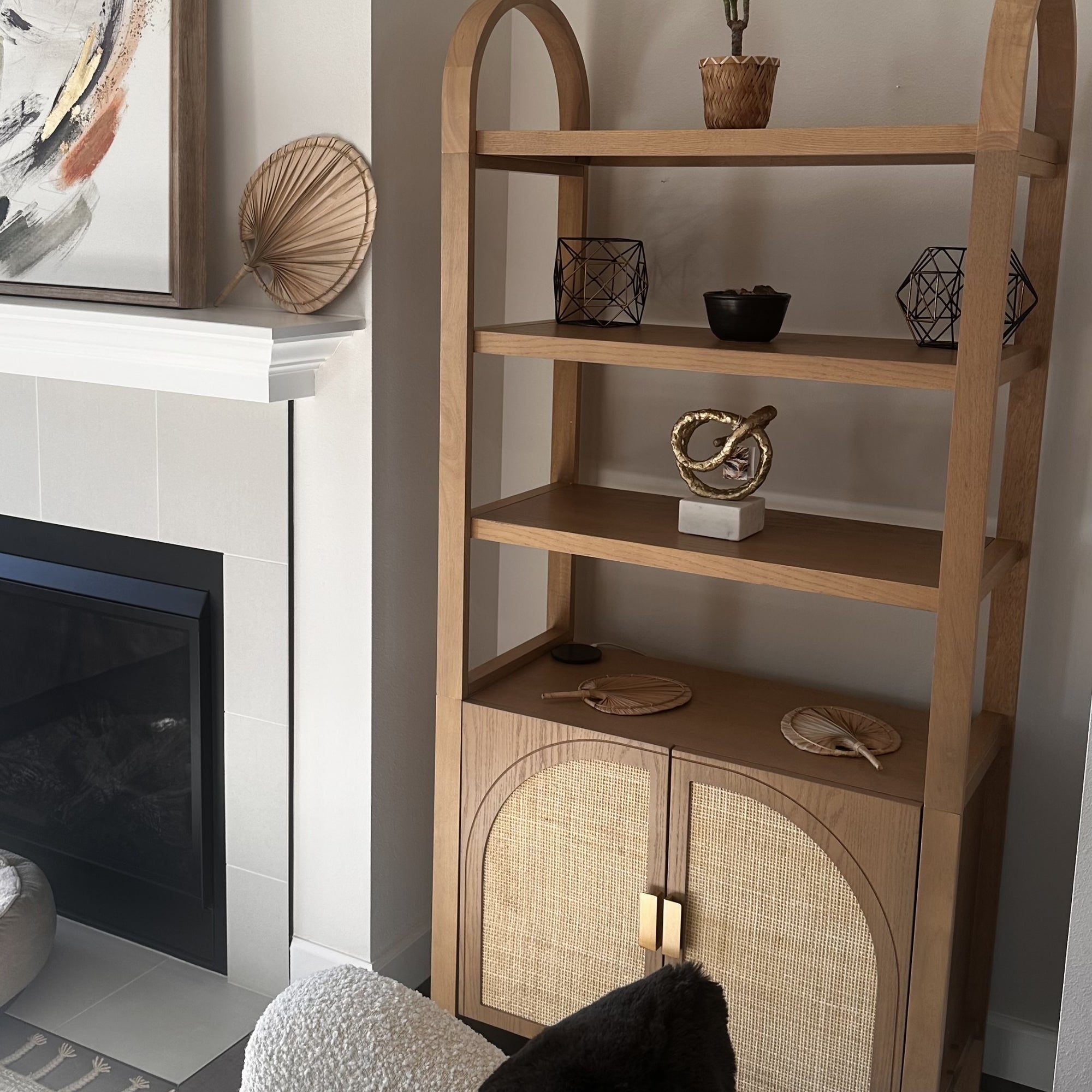 A wooden bookshelf with three shelves holds decorative objects, including geometric sculptures, a bowl, and woven fans. The lower cabinet has woven doors. The shelf is beside a fireplace and abstract wall art.