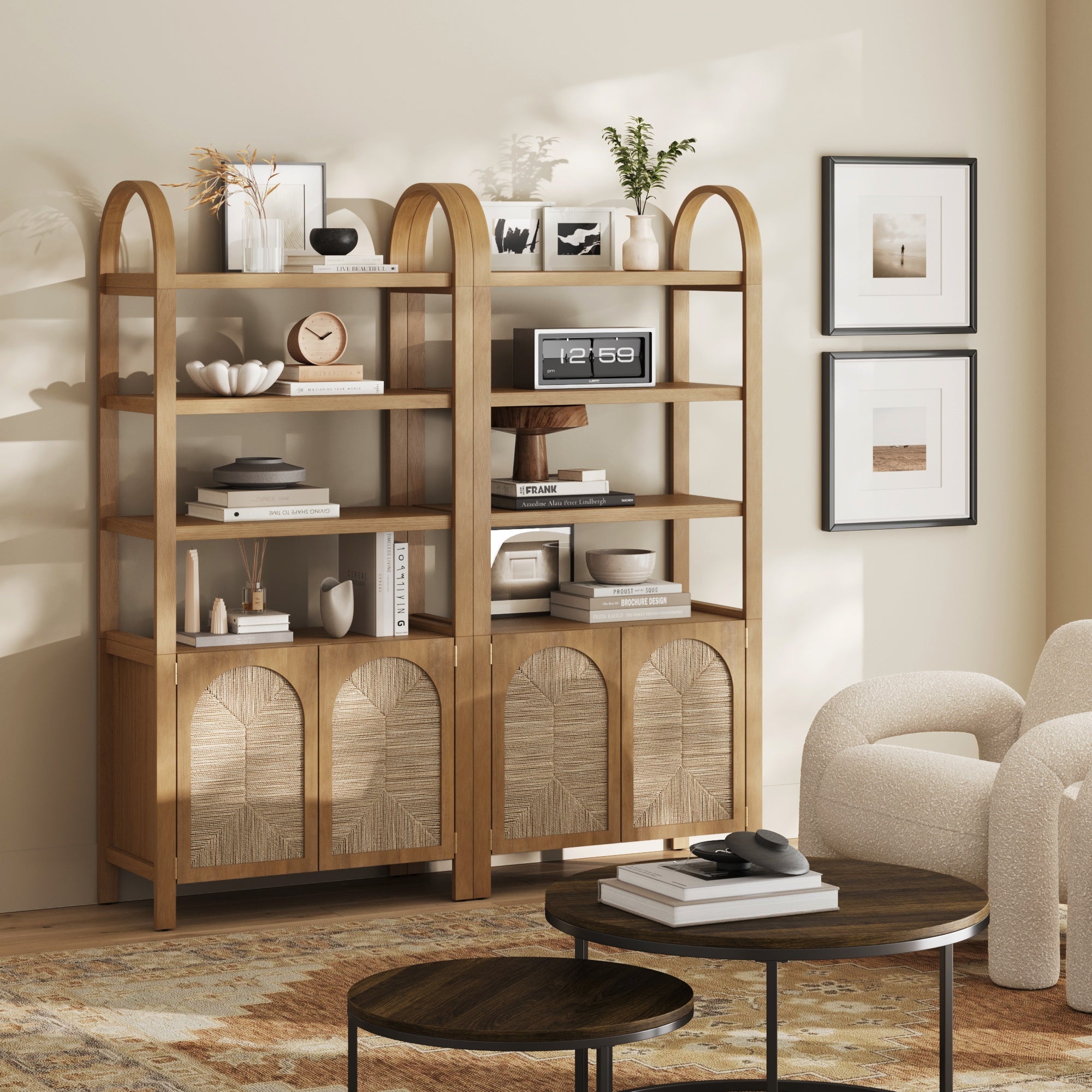 A modern living room showcases two Nathan James Boho Seagrass Arched Bookshelves in light brown with seagrass doors, styled with books and decor. Art prints hang on a cream wall; a round nested coffee table and white textured armchair sit on a rug.