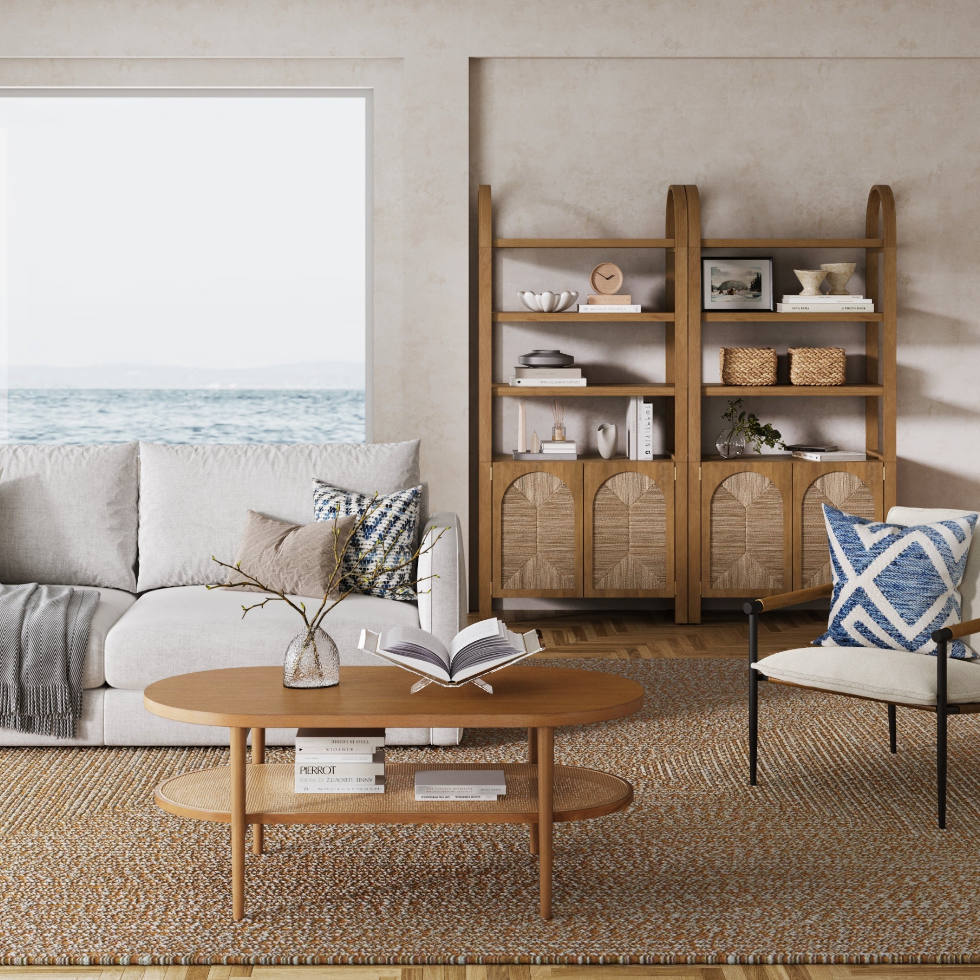 A cozy living room with a beige sofa, patterned pillows, and a wooden coffee table features a Nathan James Boho Seagrass Arched Bookshelf in light brown by the window, filled with decor and overlooking a calm ocean view.