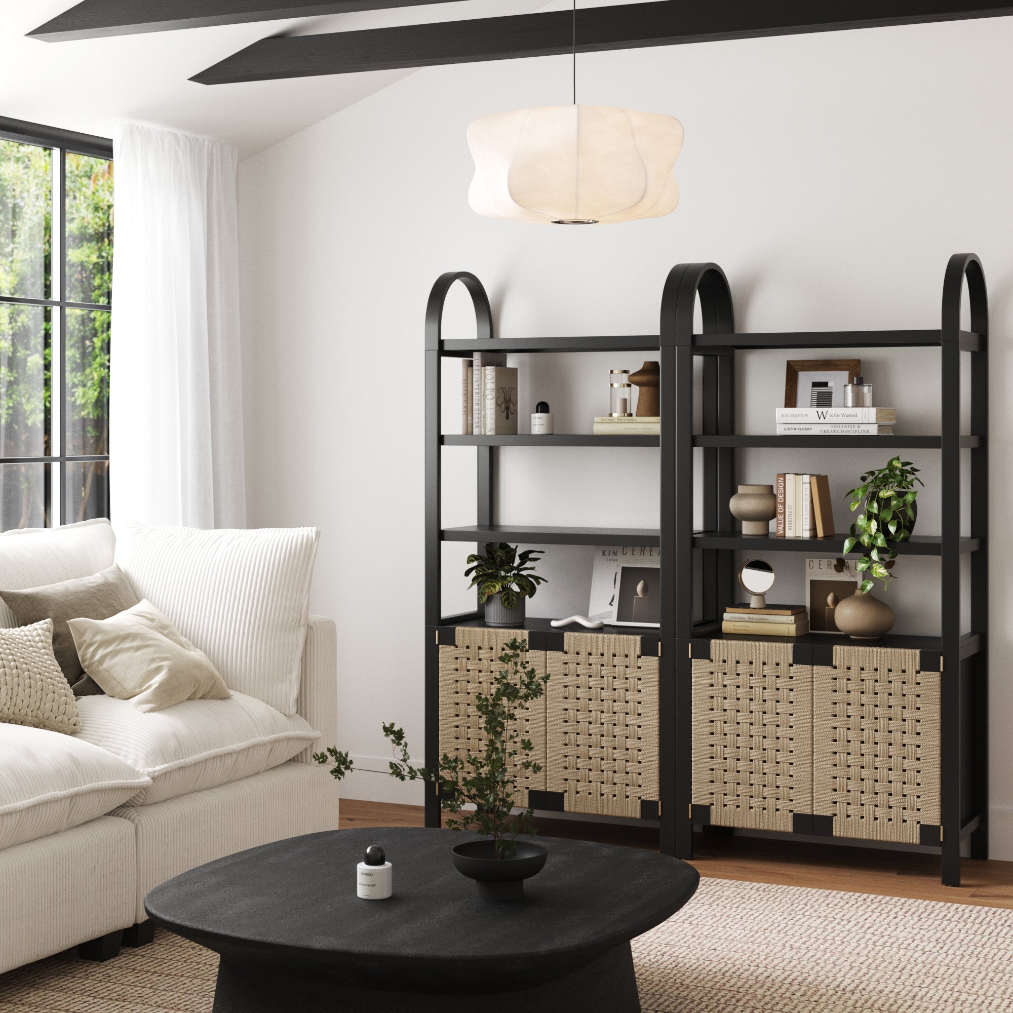 A modern living room with boho elegance highlights a white sofa, black coffee table, and Nathan James Boho Woven Seagrass Bookshelf Black (Set of 2); large windows welcome natural light, with a white pendant lamp overhead.