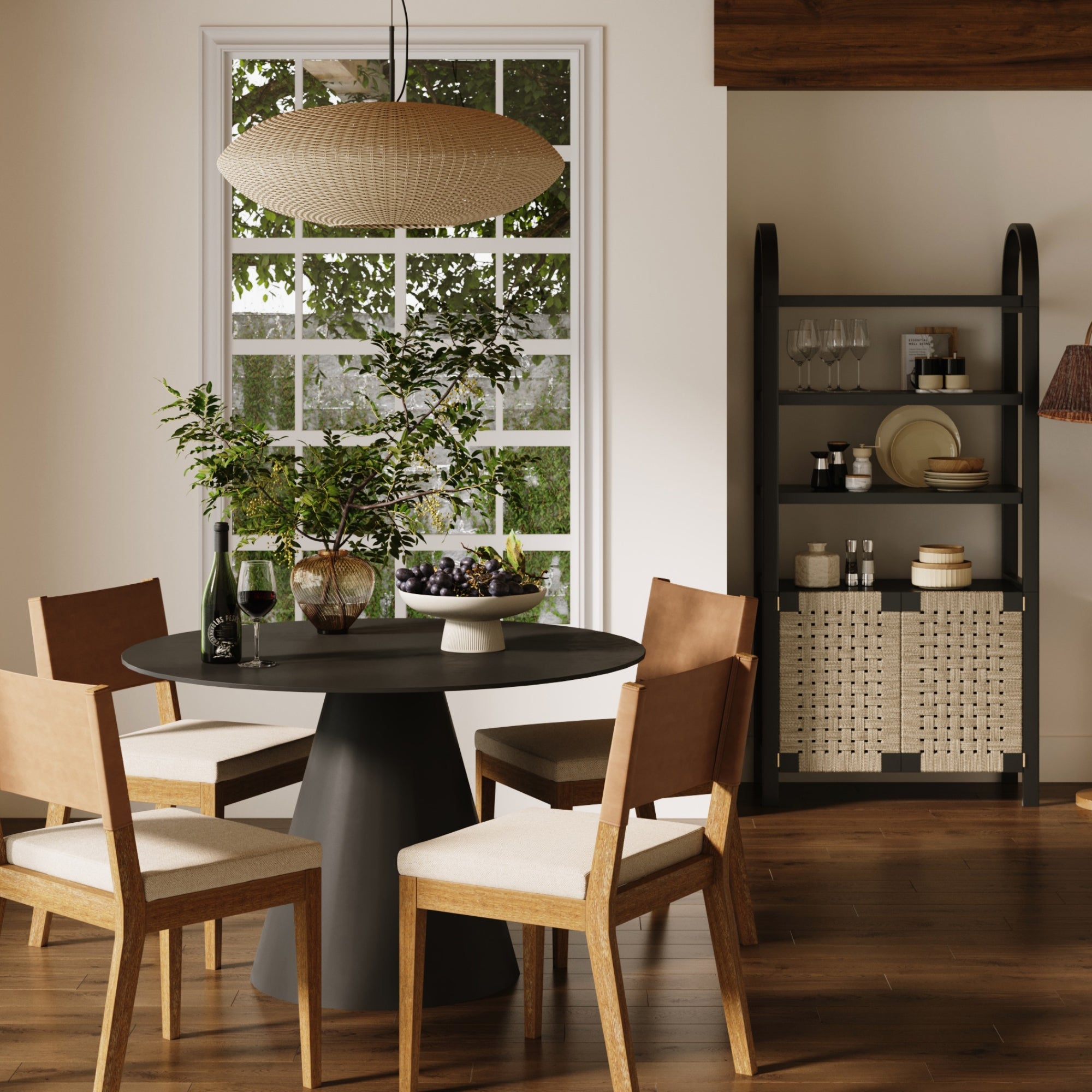 A modern dining room with a round black table, four wooden chairs, a large window, a hanging woven light fixture, and a black shelf with dishes, glasses, and woven baskets. Greenery decorates the table.
