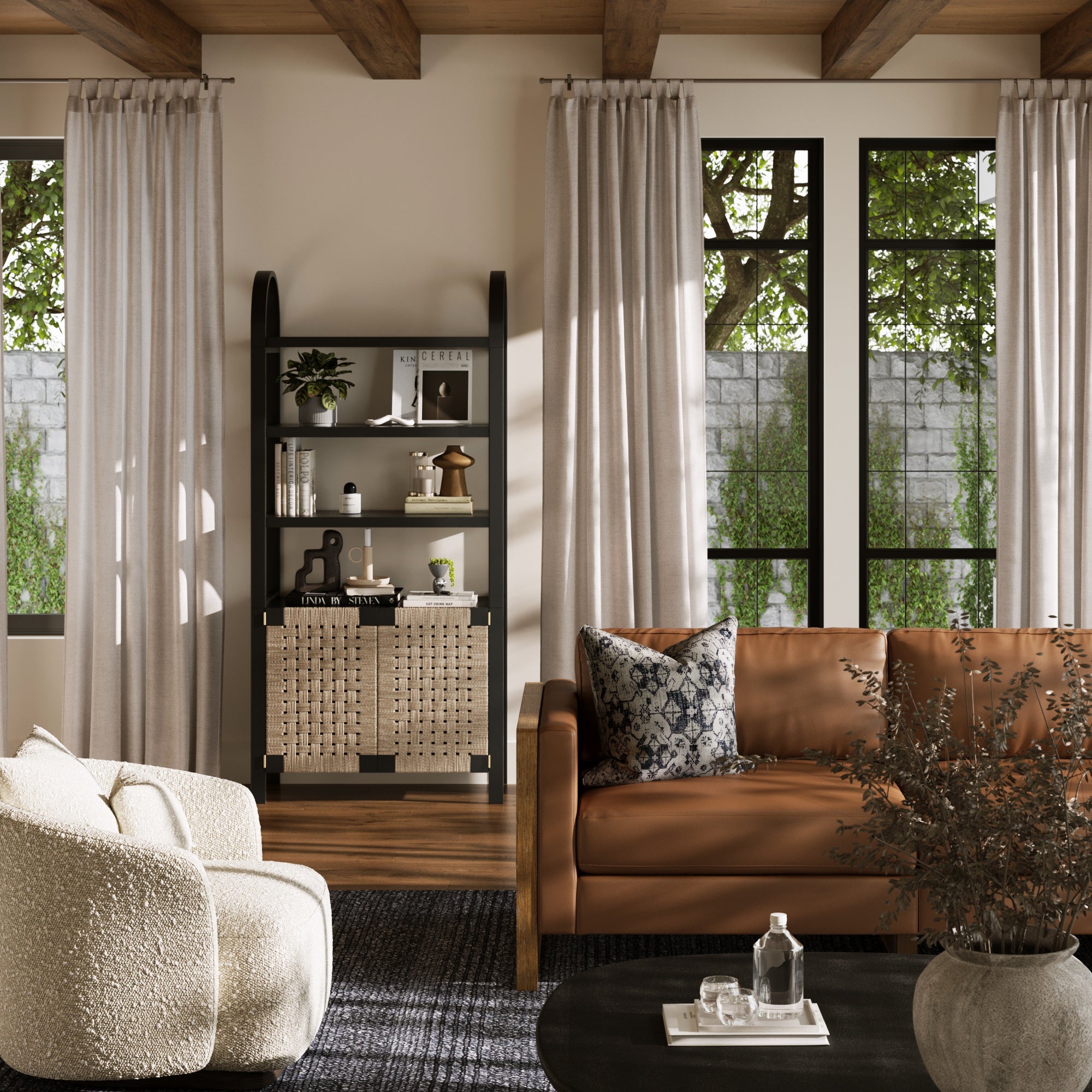 A cozy living room features a brown leather sofa, cream armchair, large windows with beige curtains, and greenery outside. The Nathan James Boho Woven Seagrass Bookshelf in black displays decor. A round black coffee table holds a vase, book, and glass items.