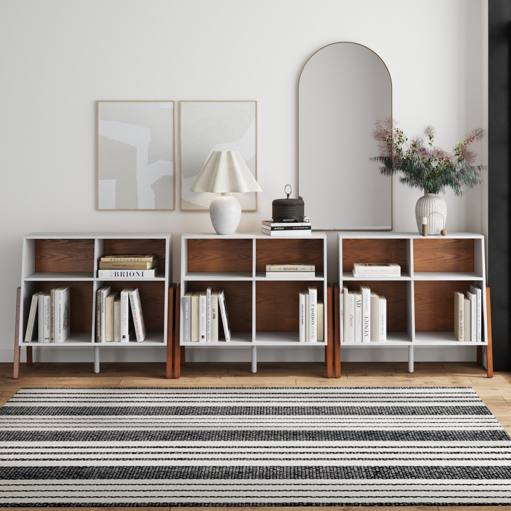 A modern living room featuring three Nathan James Large Mid-Century Open Storage Units, books, a lamp, round mirror, flower vases, abstract wall art, a Scandinavian storage cabinet, and a black-and-white striped rug on wooden floors.