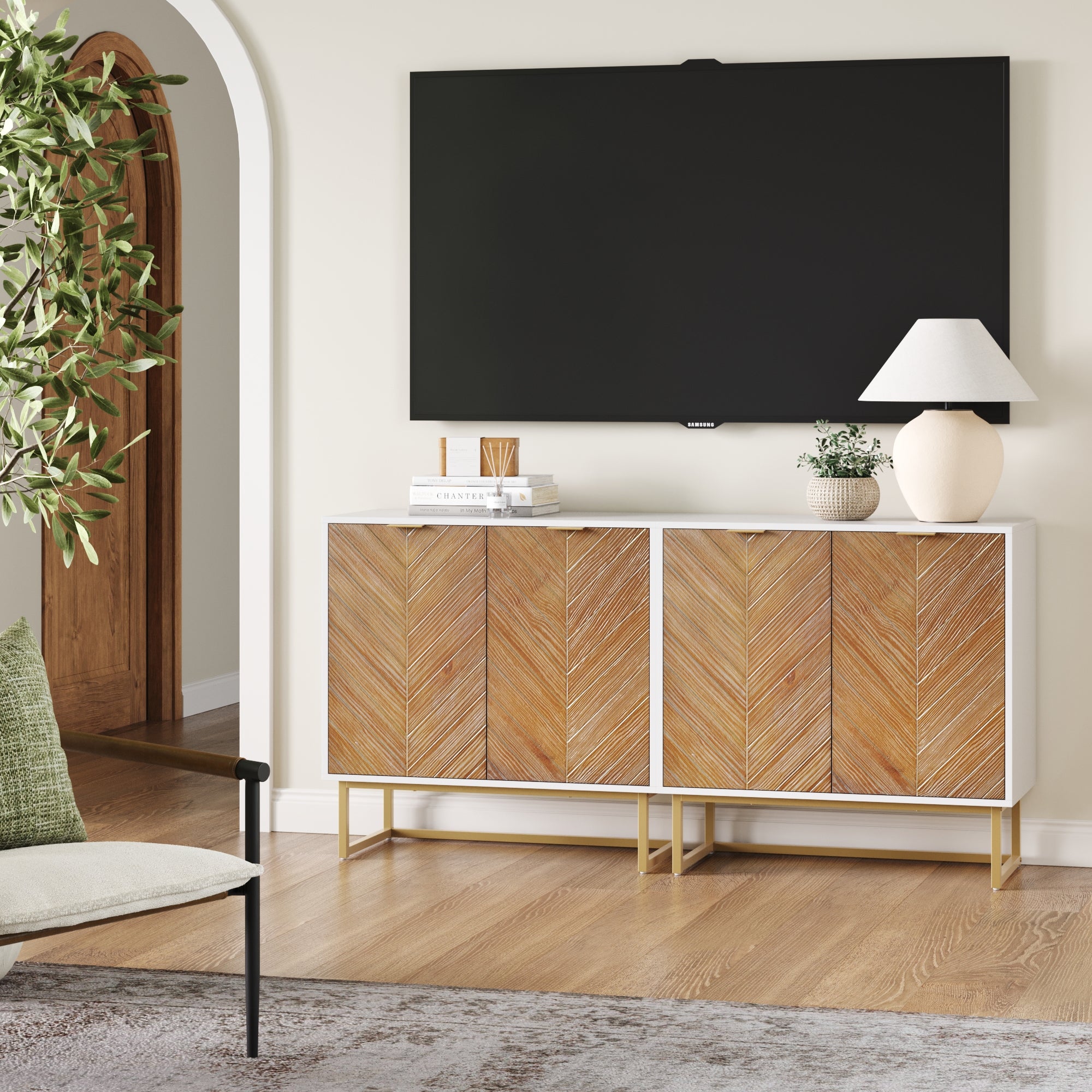 A modern living room features the Nathan James Wood Herringbone TV Console Credenza in white (set of 2), styled with a table lamp, books, vase, and decor under a wall-mounted TV, plus a potted plant and glimpses of an archway and chair.