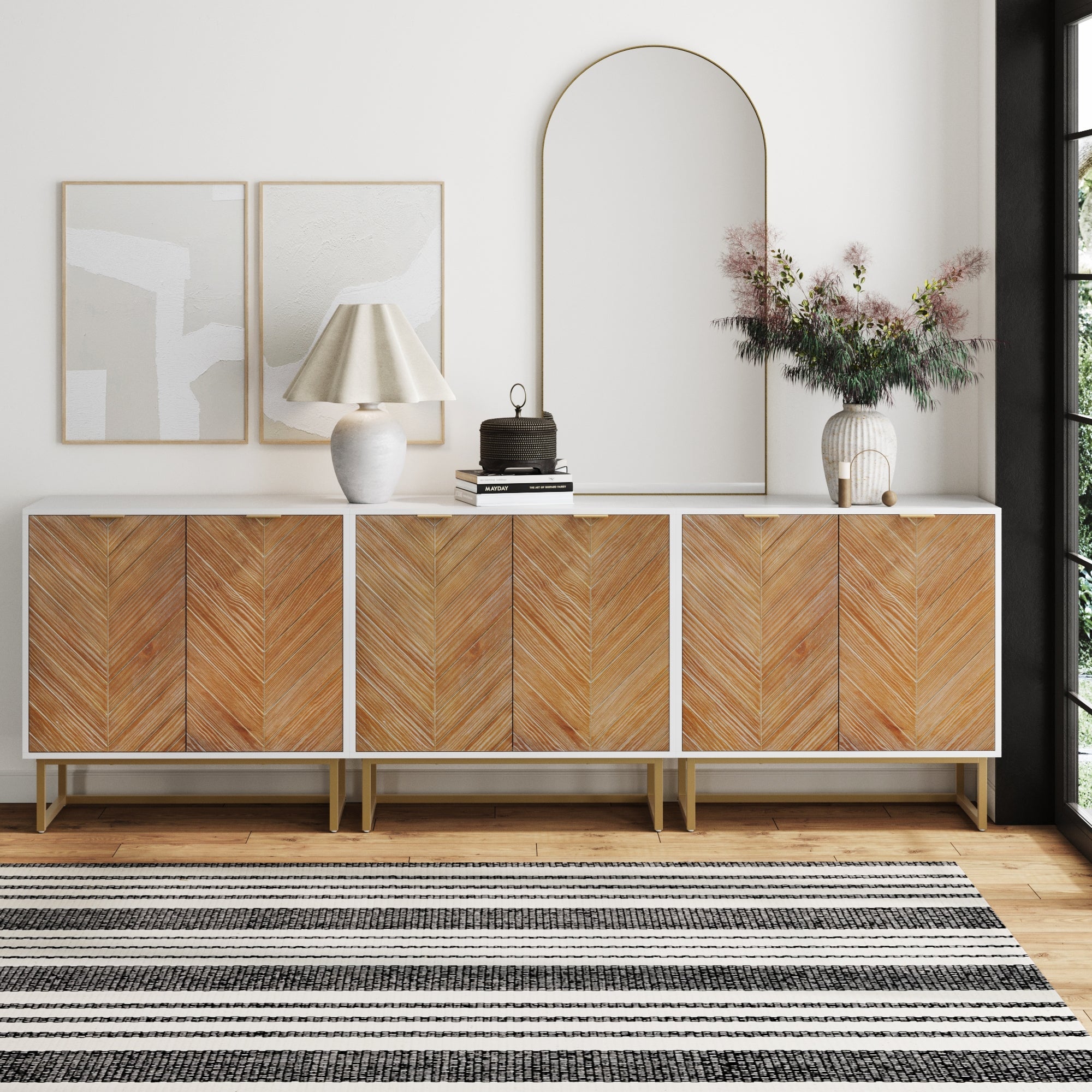 The Nathan James Herringbone TV Console Credenza White (Set of 3) features chevron wood doors and sits against a white wall, styled with a lamp, books, vases, and an arch mirror. Abstract art hangs above; a black-and-white striped rug completes the look.