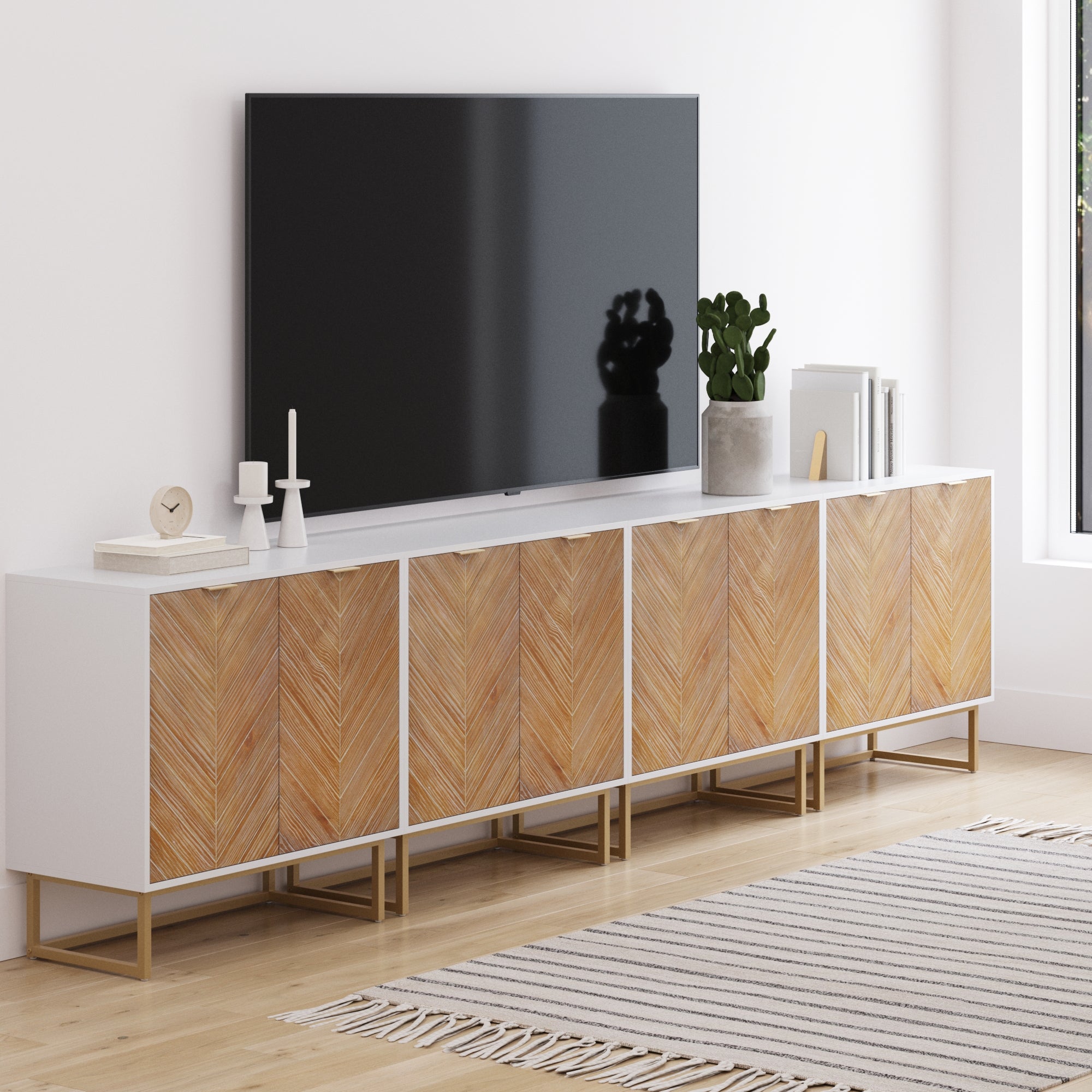 A modern living room with a Large Herringbone TV Console Credenza White (Set of 4) by Nathan James, displaying a flat-screen TV, plants, books, and decor beside a window with a striped rug on light wood floors.