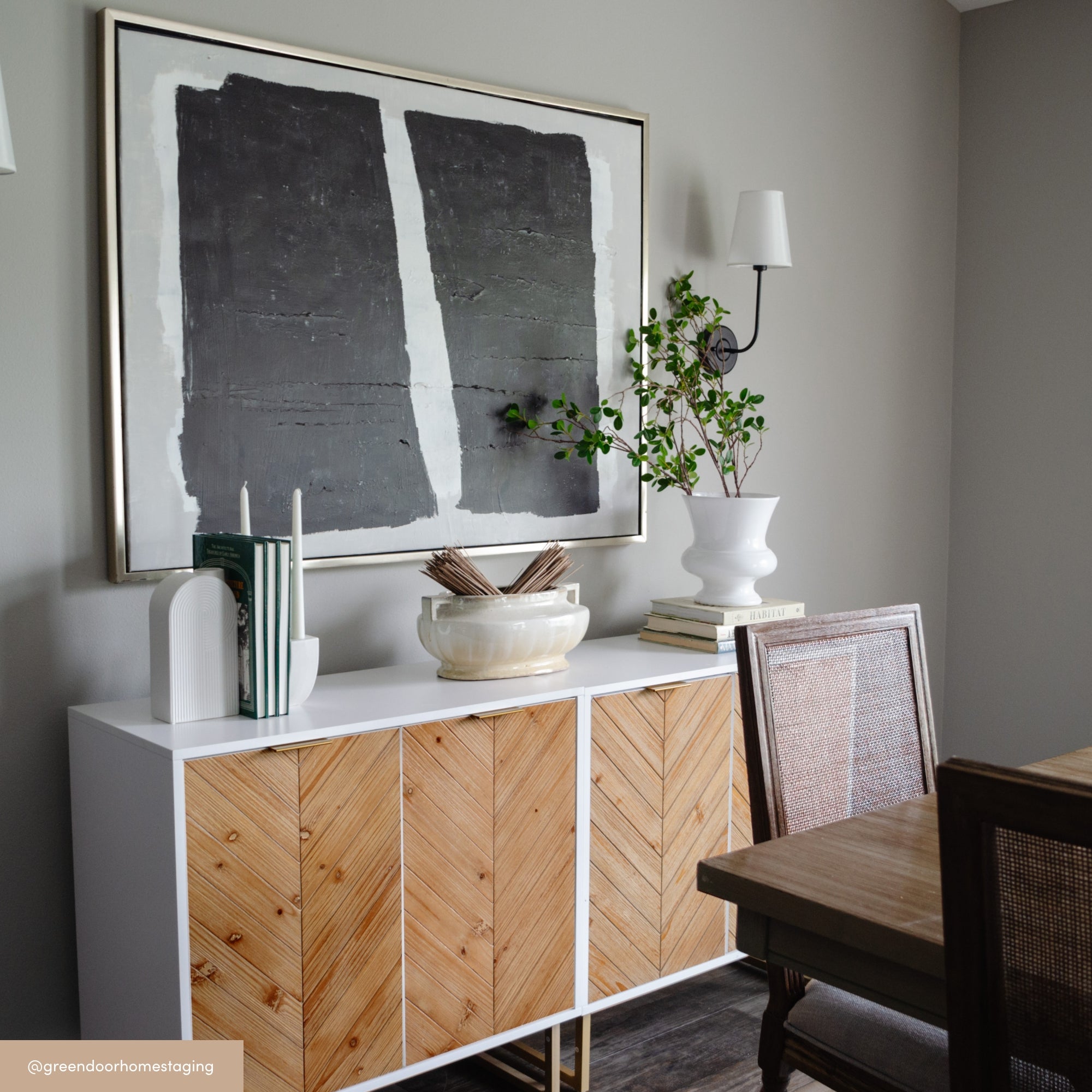 A modern dining room with a white and wood sideboard, abstract black and gray wall art, a white vase with greenery, decorative books, and wooden chairs with cane backs. The room has neutral-colored walls.
