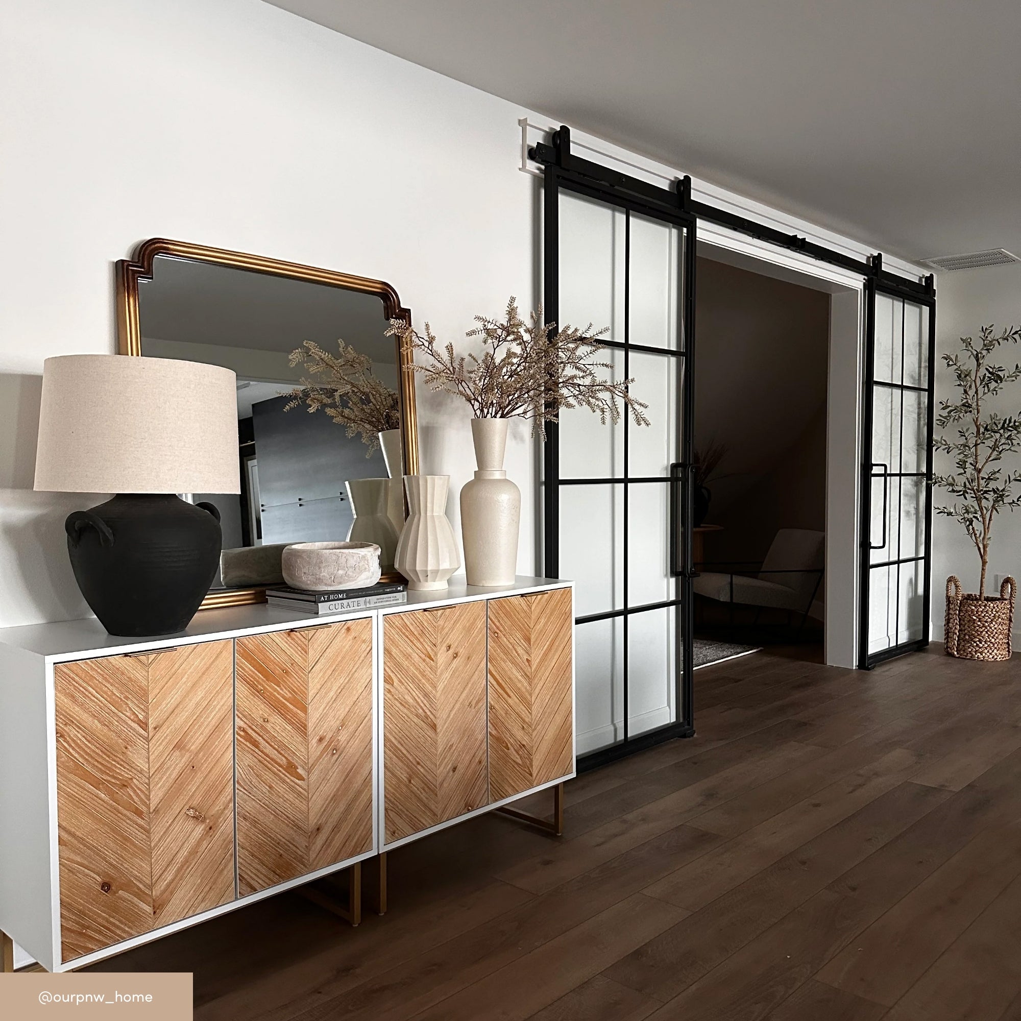 A modern entryway with a white and wood chevron cabinet, topped with a black lamp, stacked books, and decorative vases. A gold-framed mirror is above the cabinet, and black-framed glass sliding doors are in the background.