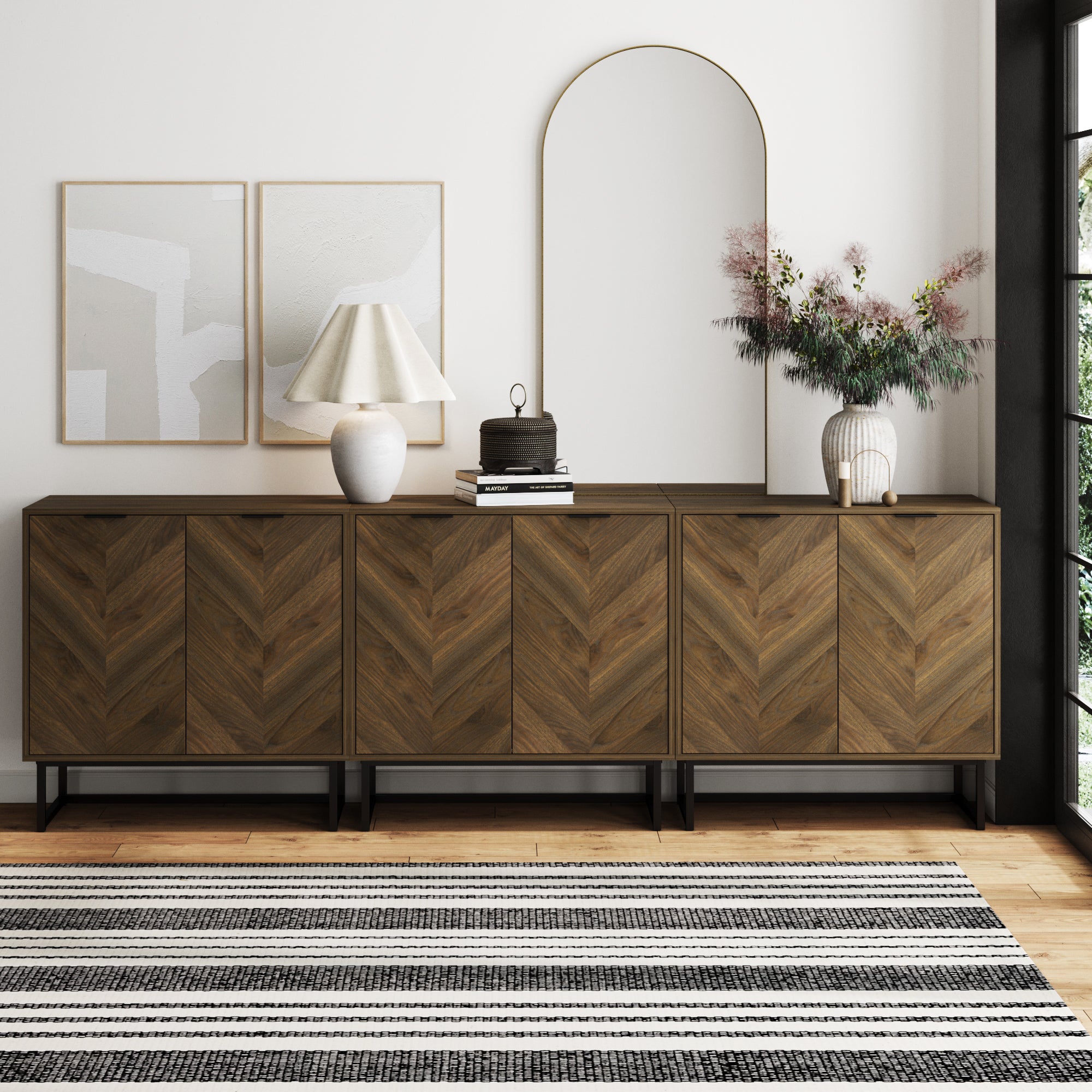 A modern living room features a Nathan James Herringbone TV Console Credenza Walnut (Set of 3), two abstract art pieces, a table lamp, stacked books, a mirror, a white vase with pink flowers, and a black-and-white striped rug.