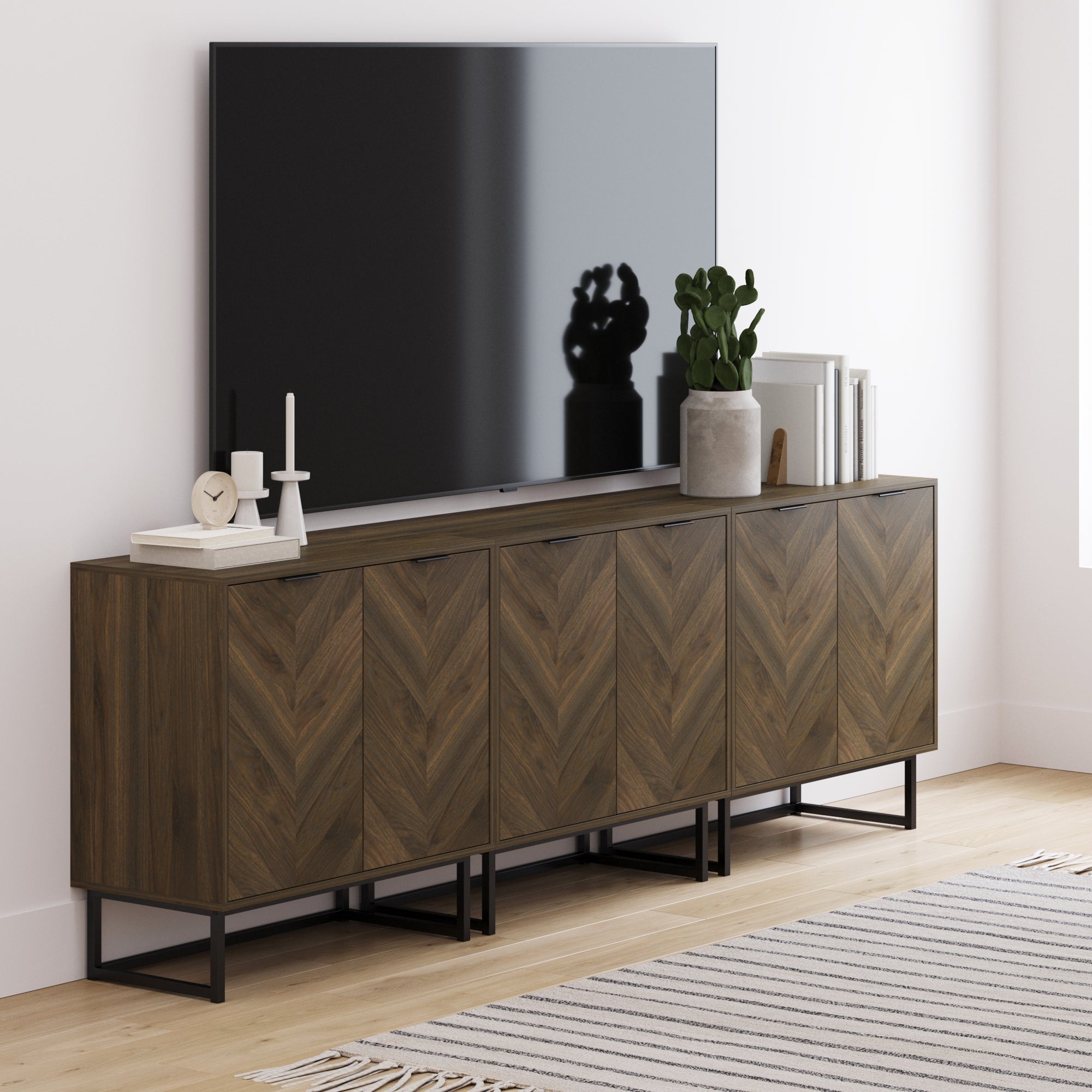 A modern living room features a large flat-screen TV on the Nathan James Herringbone TV Console Credenza Walnut (Set of 3), styled with books, a plant, candles, and decor. A striped rug covers the light wood floor.