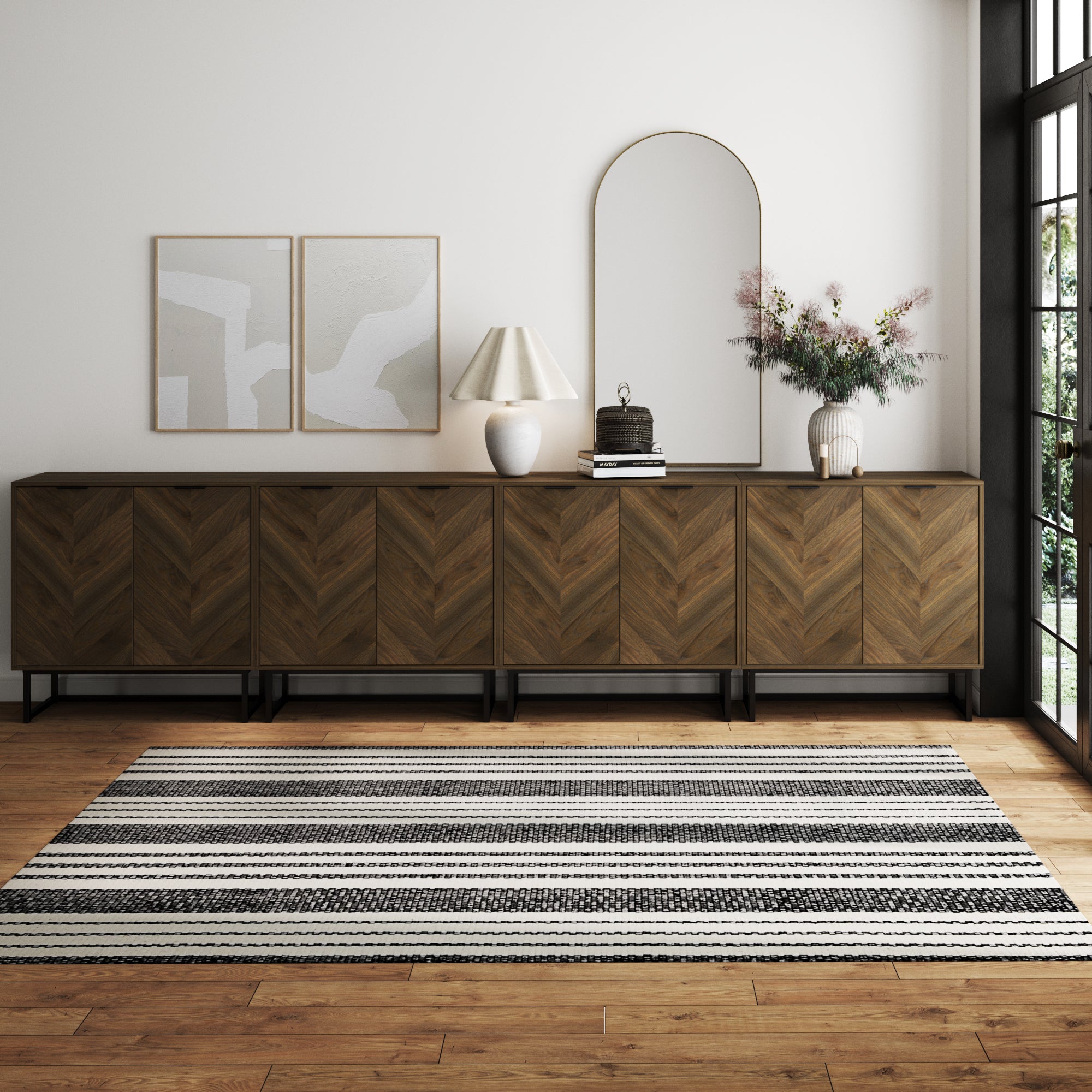 Modern living room with Nathan James Large Herringbone Credenza Console Table in walnut (set of 4), a striped rug, two abstract art pieces, table lamp, decorative books, and a vase with flowers by sunlit windows.