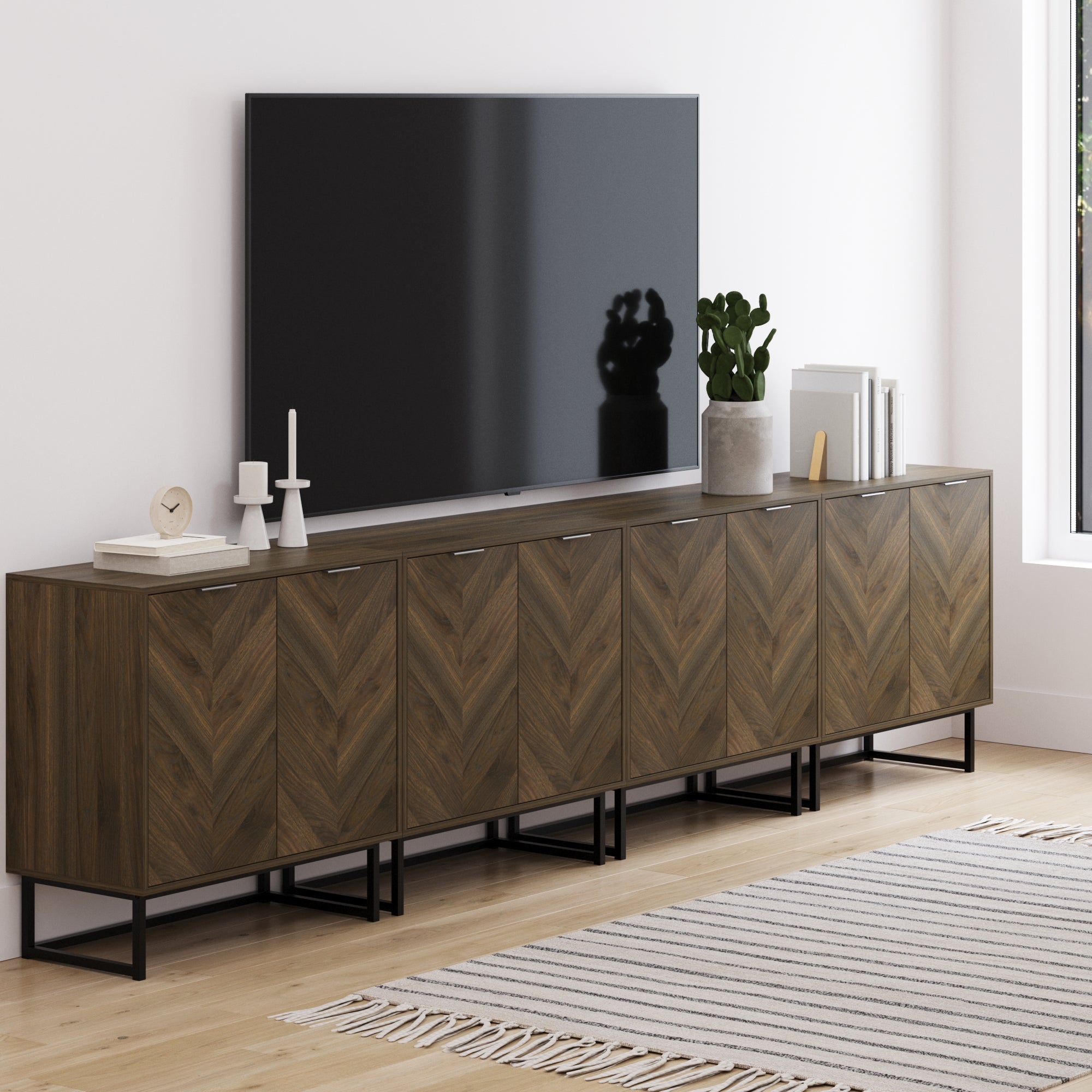A modern living room featuring the Nathan James Large Herringbone Credenza Console Table in Walnut (Set of 4) with décor, books, and a plant on top, positioned beside a patterned rug on a light wood floor with a large TV above.