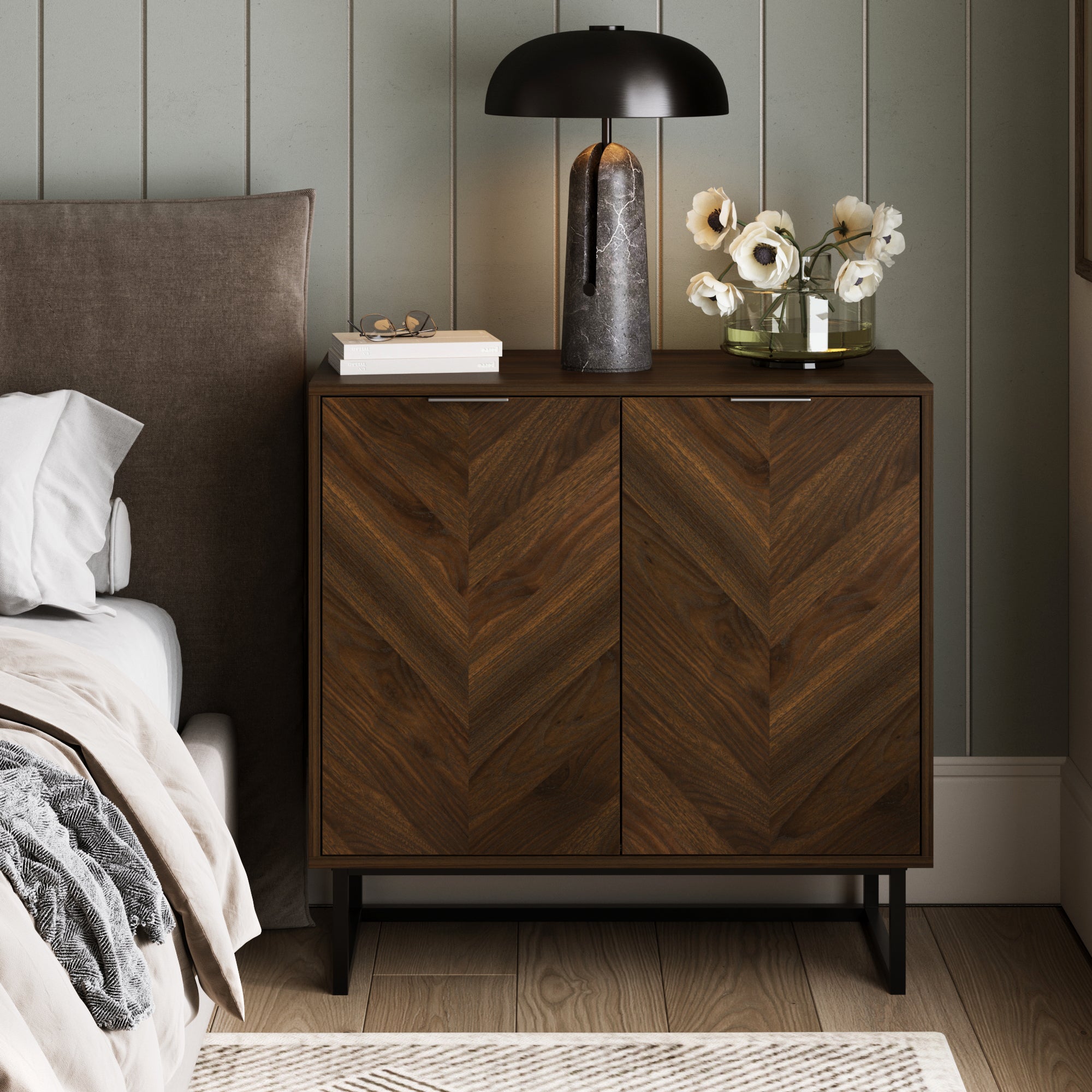A Nathan James Wood 2-Door Storage Cabinet in Walnut with a herringbone pattern stands by the bed. On top are a black lamp, glass vase with white flowers, and books. The background features vertical wall paneling.