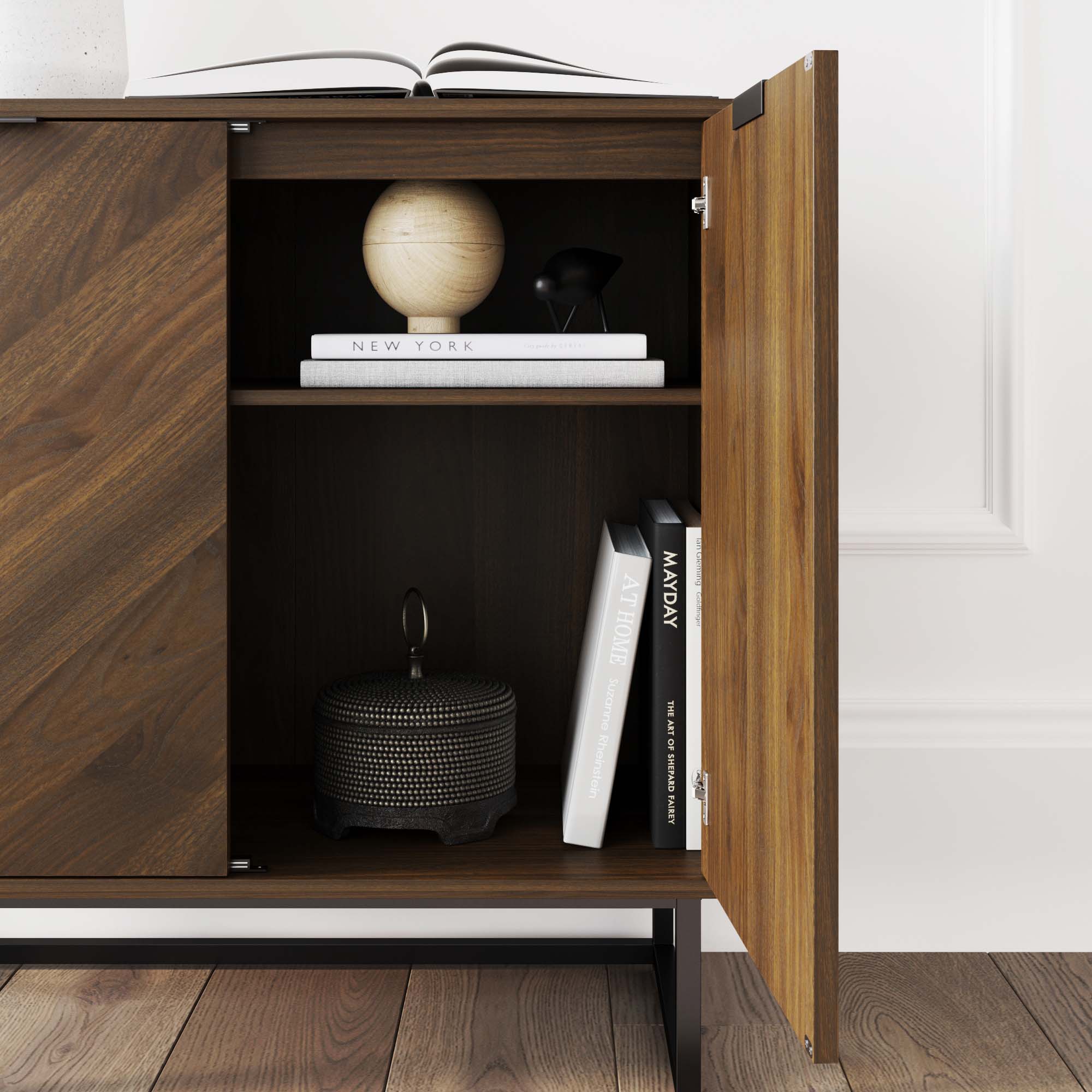 A wooden cabinet with one door open, revealing a shelf with books, a round wooden decor piece, a small black figurine, and a textured dark container with a handle. An open book rests on top of the cabinet.