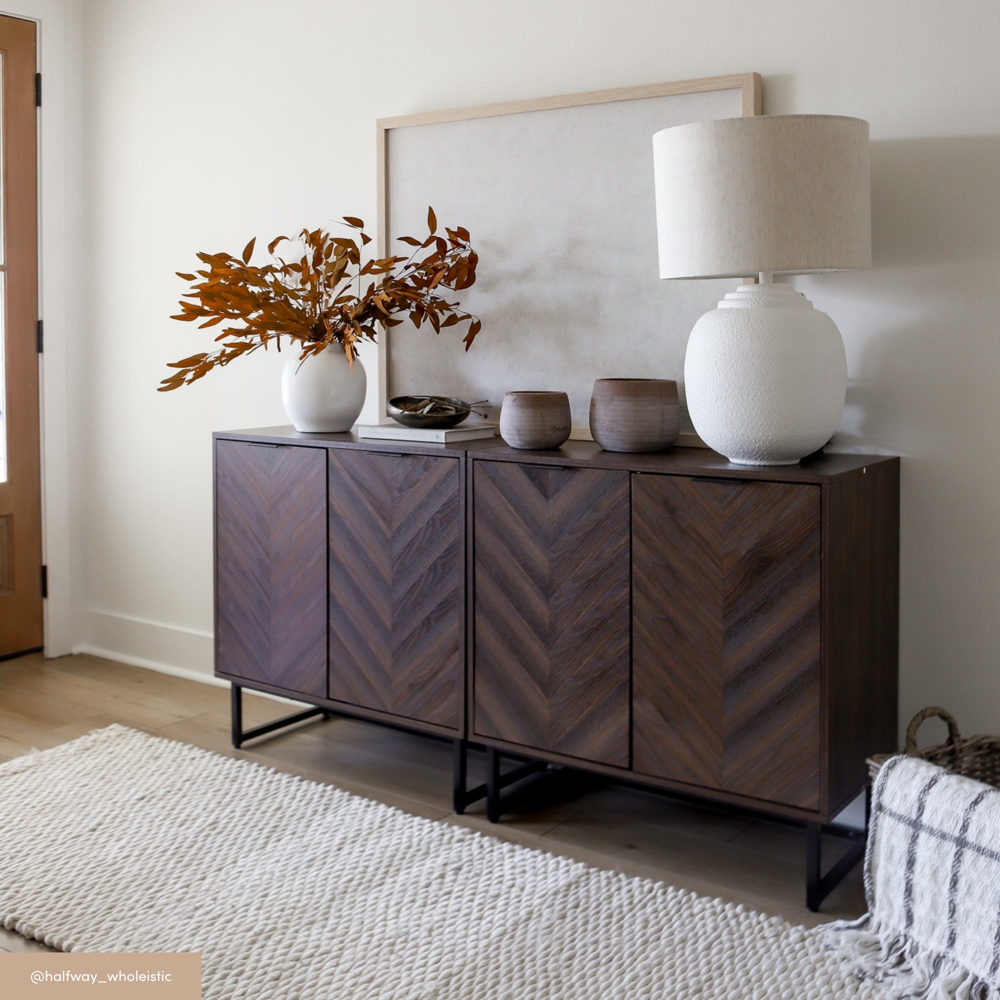 A modern wood console table with a herringbone pattern holds a white textured lamp, ceramic vases, and a vase with brown foliage. A neutral rug and a framed picture complete the minimalistic decor.