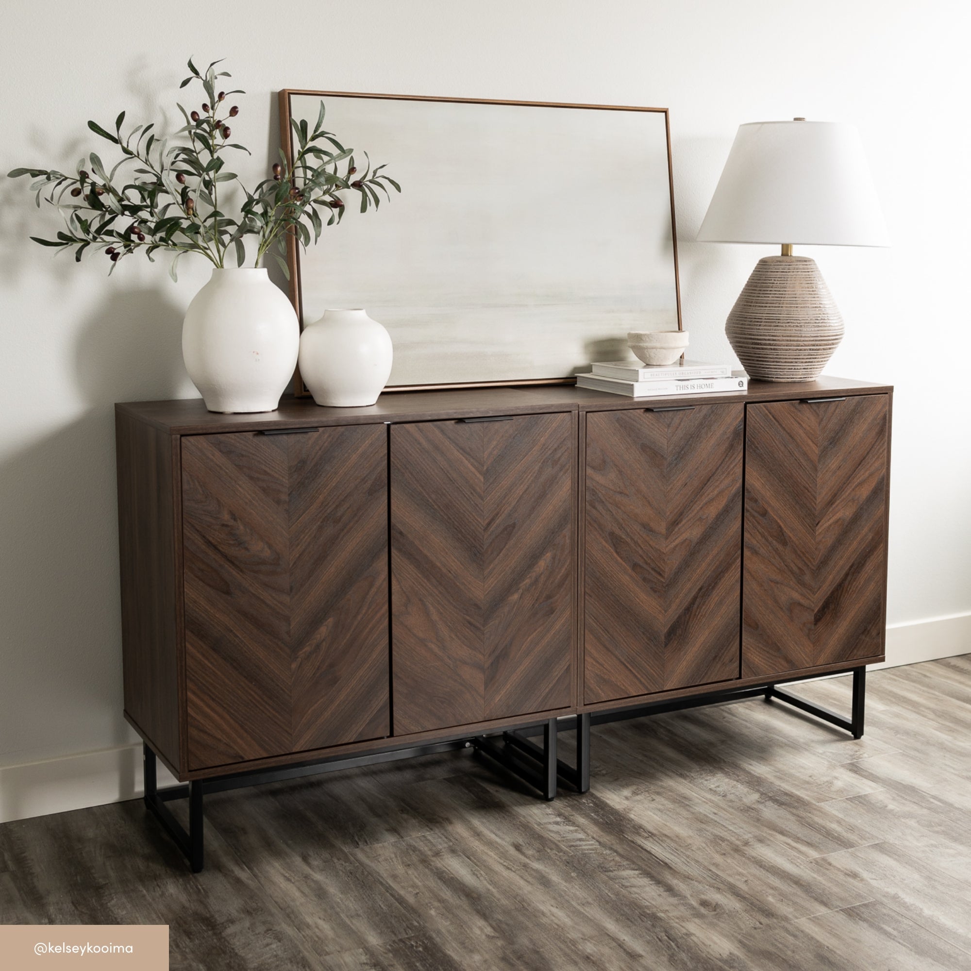 A modern wooden sideboard with a chevron pattern stands against a white wall, topped with white vases holding greenery, a framed abstract artwork, and a textured table lamp. The floor is light wood.