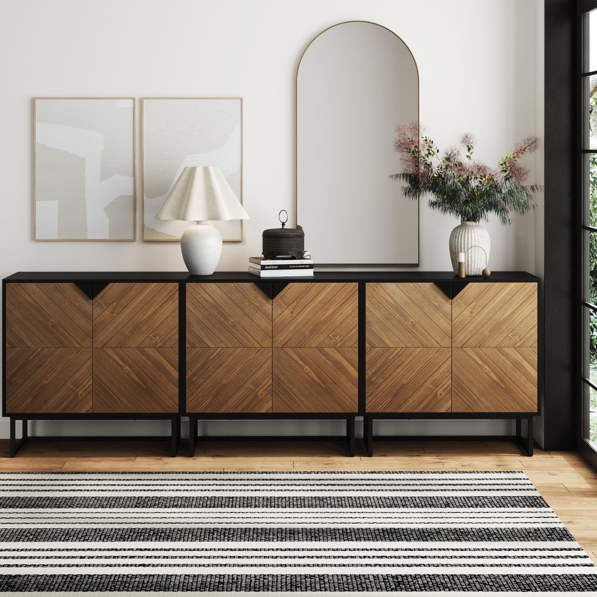 The Nathan James Brushed Fir TV Console Credenza (Set of 3) displays a lamp, books, jar, and vase with foliage. Above it hang two abstract art prints and an arched mirror. A striped rug lies on the floor.