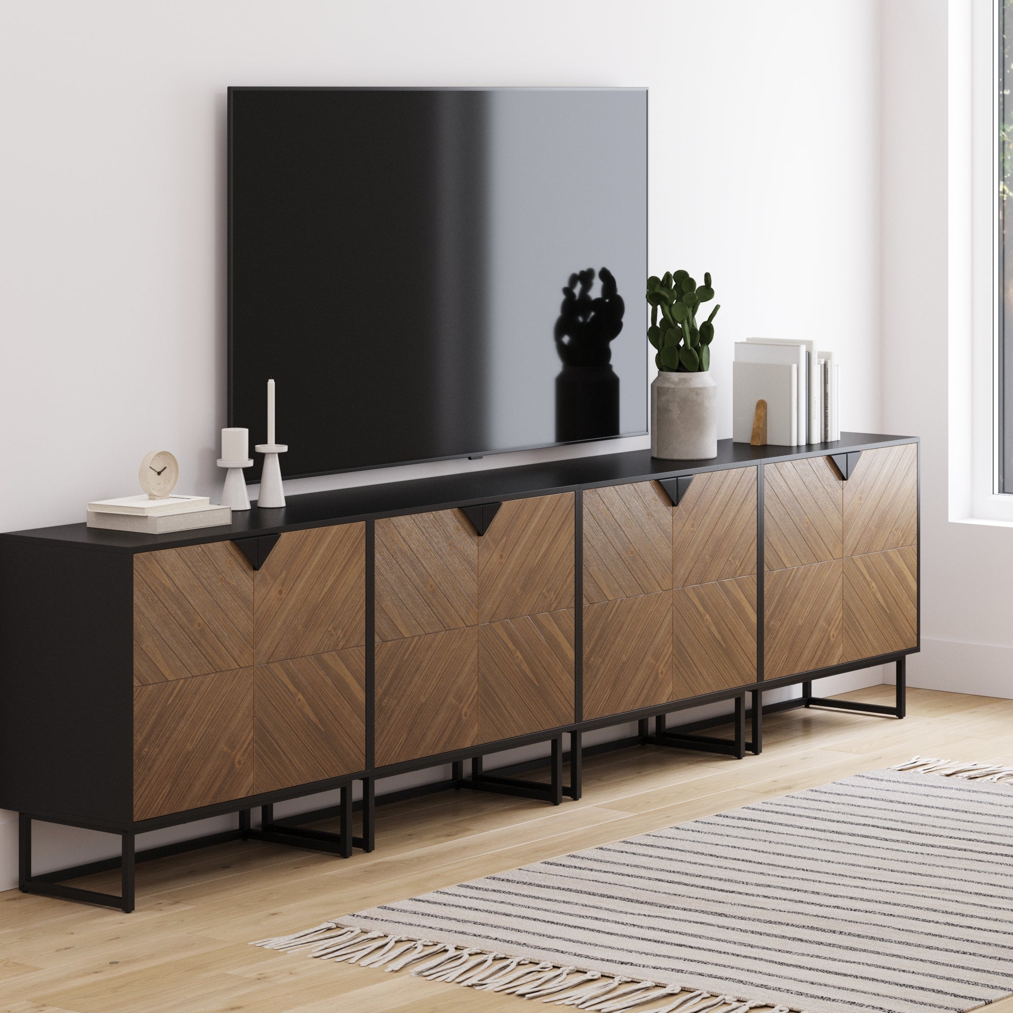 A modern living room features a large TV on the Nathan James Large Brushed Fir Credenza Console Table (Set of 4), decorated with candles, a plant, books, and accents. A striped rug and sunlit window complete the look.