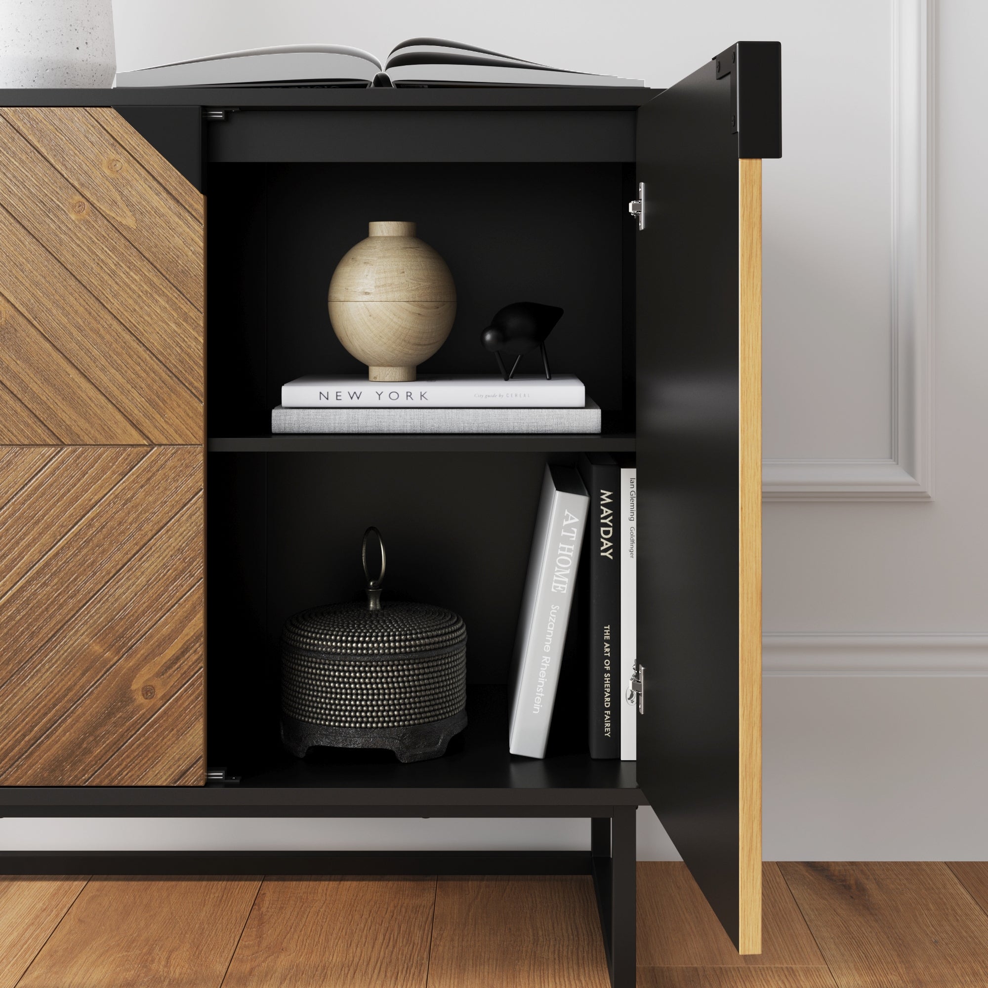 A modern black cabinet with wooden accents sits on a wood floor. Its door is open, revealing shelves with a ceramic vase, books, a decorative black object, and a round, textured container. An open book rests on top.