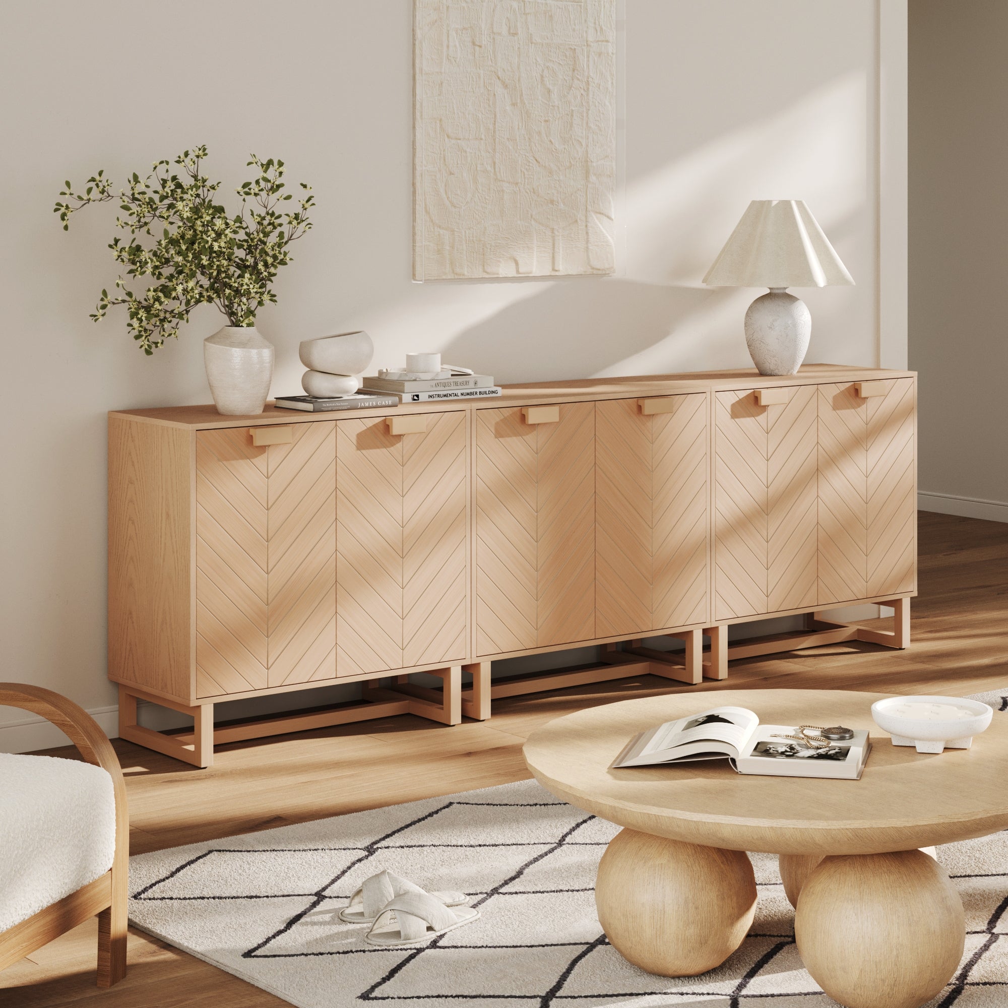 A modern living room features three Nathan James Wood Herringbone 2-Door Accent Cabinets in Raw Oak, decorative vases, a lamp, a small plant, abstract wall art, a round wooden coffee table, and a geometric rug on a wooden floor with sunlight streaming in.