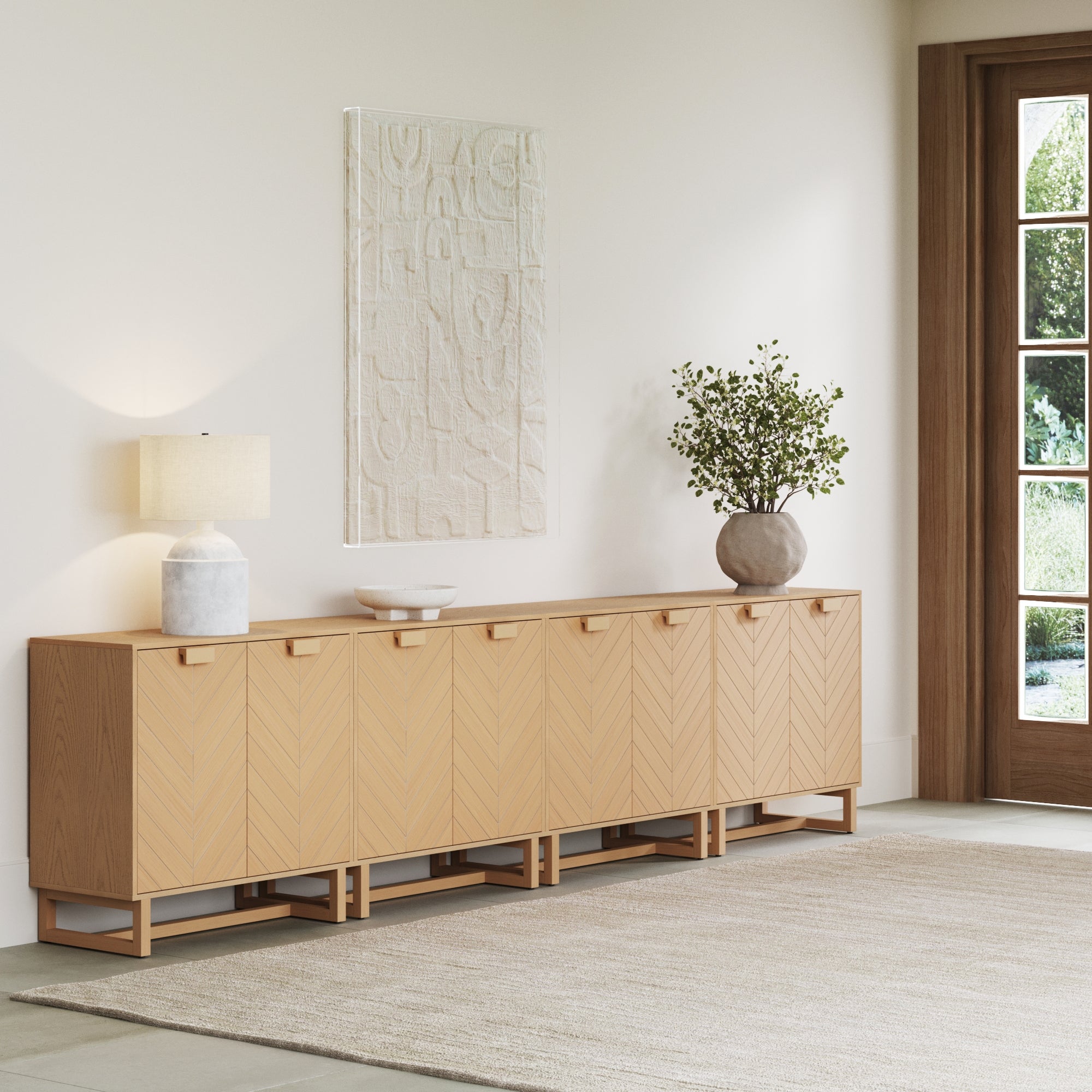The Nathan James Wood Herringbone 2-Door Credenza in Raw Oak (Set of 4) features chevron-patterned doors and sits against a white wall with a lamp, a white vase holding green branches, and abstract wall art; a large window and beige rug are seen nearby.