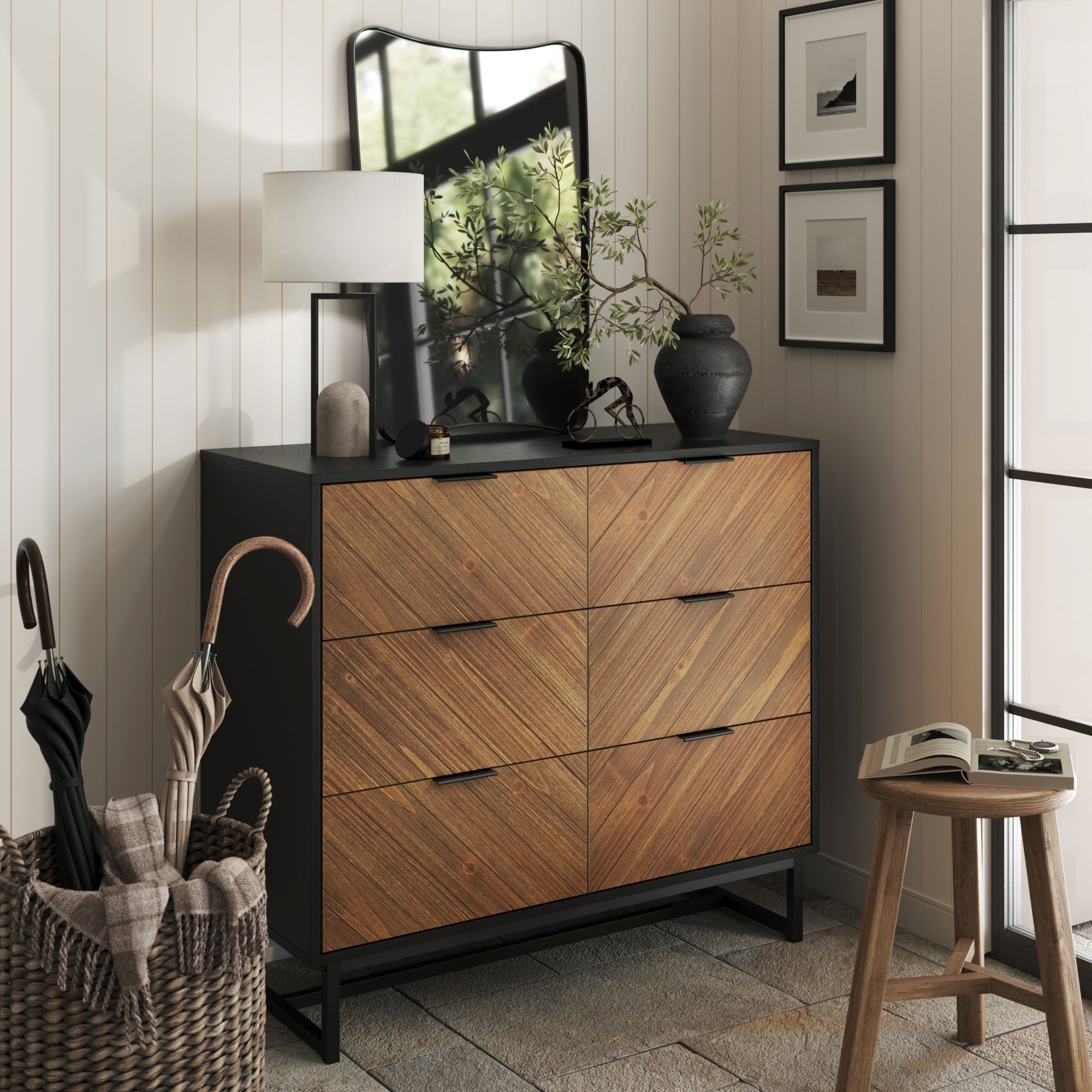 A modern entryway features the Nathan James 3-Drawer Brushed Fir Dresser with a black frame and wood drawers, topped with a lamp, mirror, vases, and a branch. Nearby are framed art, a wooden stool, and a woven basket holding umbrellas.