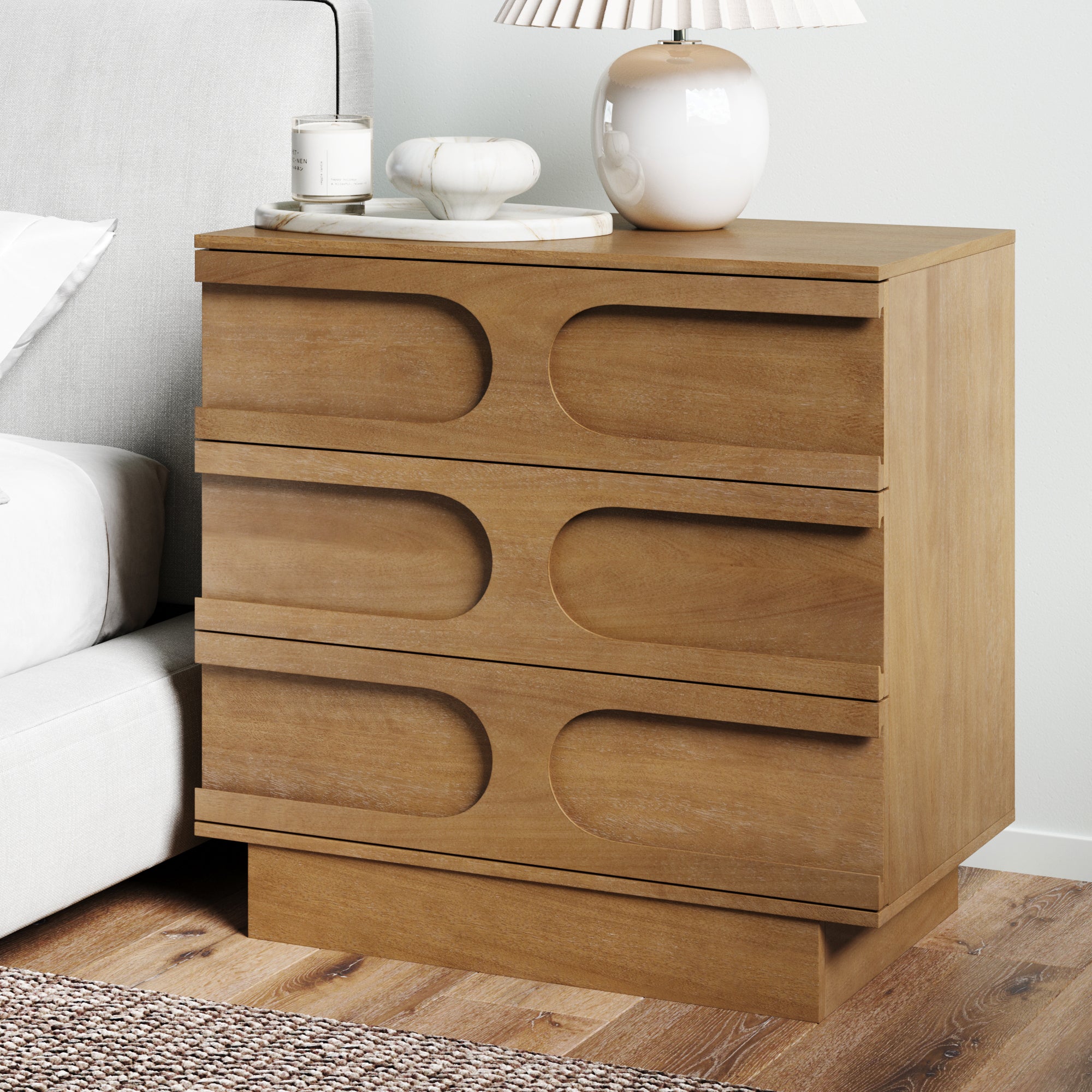 A wooden nightstand with three drawers featuring curved, geometric designs, topped with a white table lamp, a candle, and decorative items. The nightstand is next to a bed with white bedding.