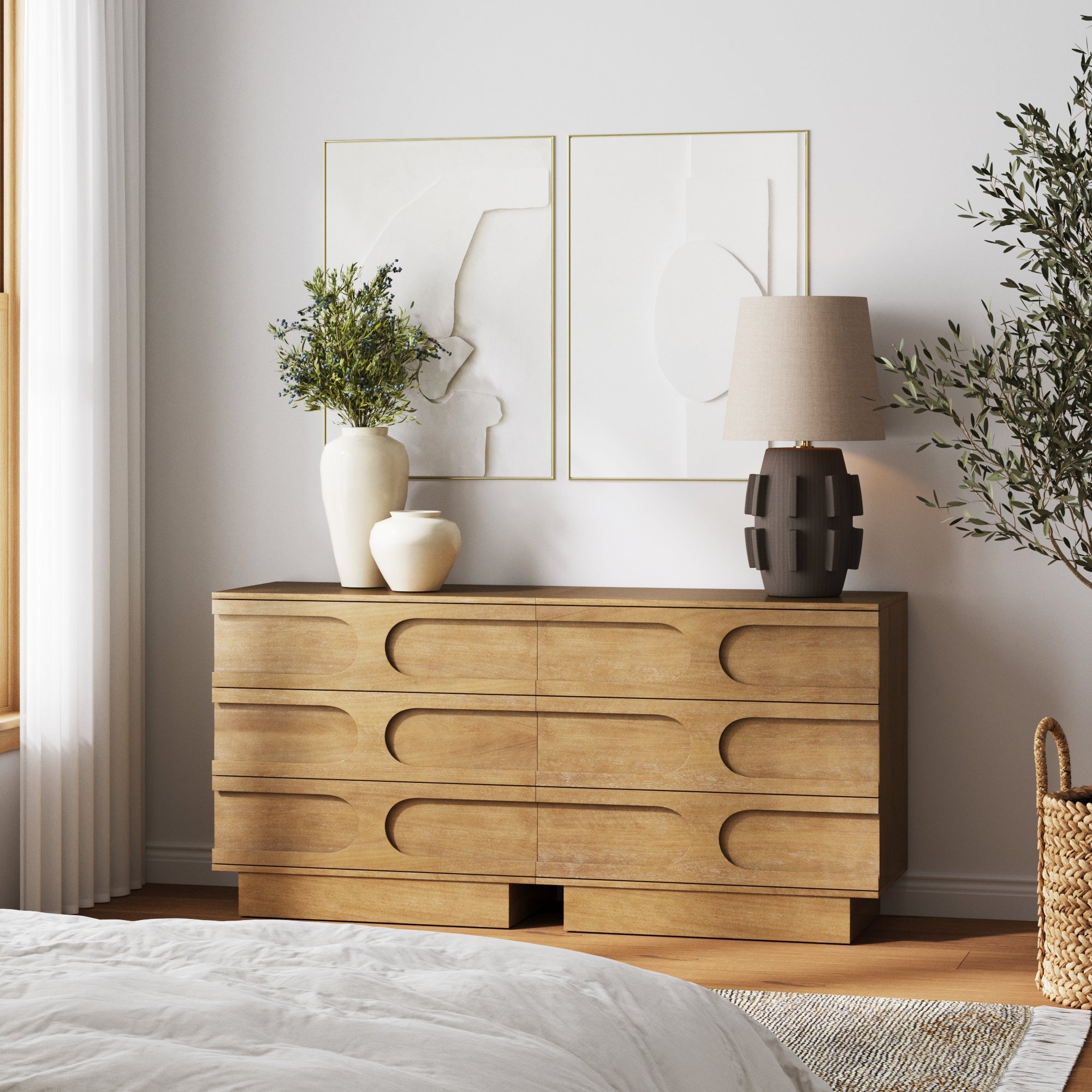The Nathan James Mid-Century 3-Drawer Dresser in Light Brown sits against a white wall, topped with two vases, a table lamp, and framed abstract art. Nearby are a plant and woven basket, with part of a bed visible in the foreground.