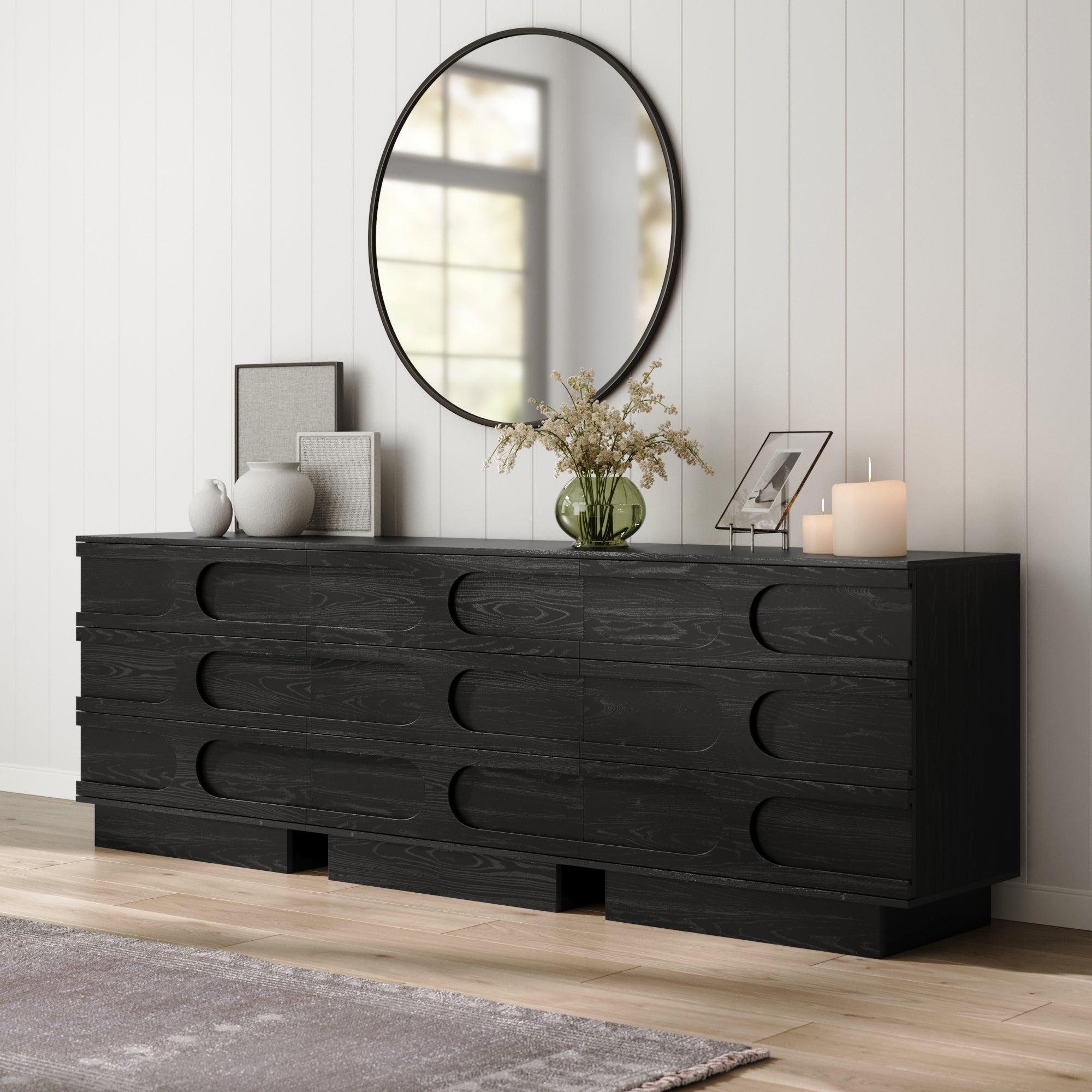 The Nathan James Mid-Century 3-Drawer Arched Dresser in Black Oak (Set of 3) features unique handles, topped with a round mirror, framed photos, vases, flowers in a green glass vase, and candles. White paneled walls and light wood floors complete the look.