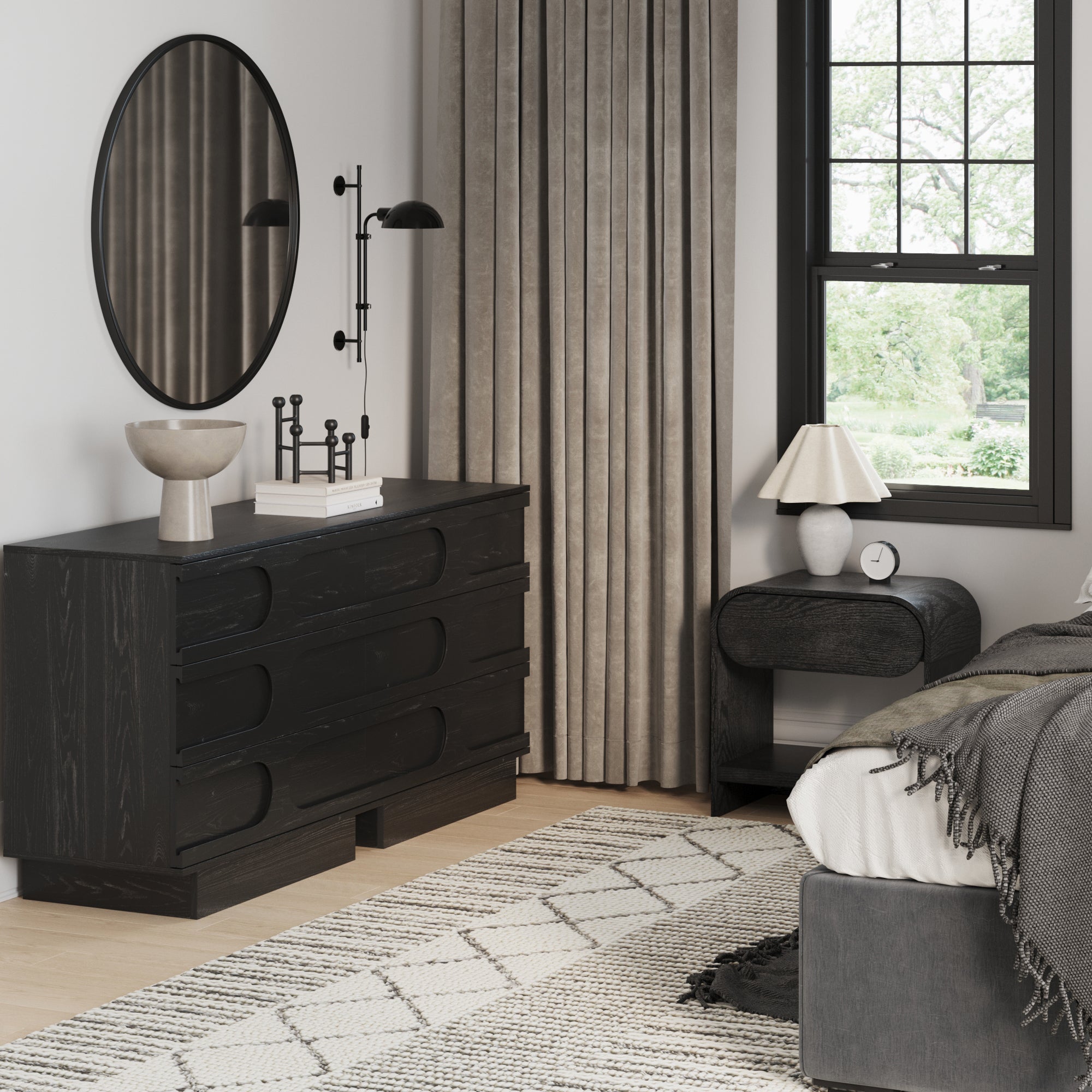 A modern bedroom showcases the Nathan James Mid-Century Two Arched Dressers and Nightstands Set in Black Oak, paired with a round mirror, lamp, books, decor pieces, gray bedding, and large windows with neutral curtains letting in natural light.