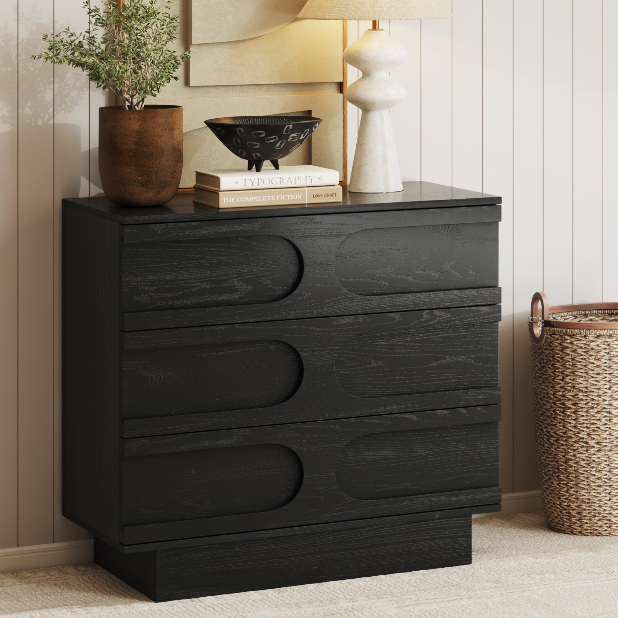 The Nathan James Mid-Century 3-Drawer Arched Dresser in Black Oak features curved drawer fronts and is styled with a potted plant, two books, a bowl, and a lamp. A woven basket sits on the floor beside this elegant storage piece.