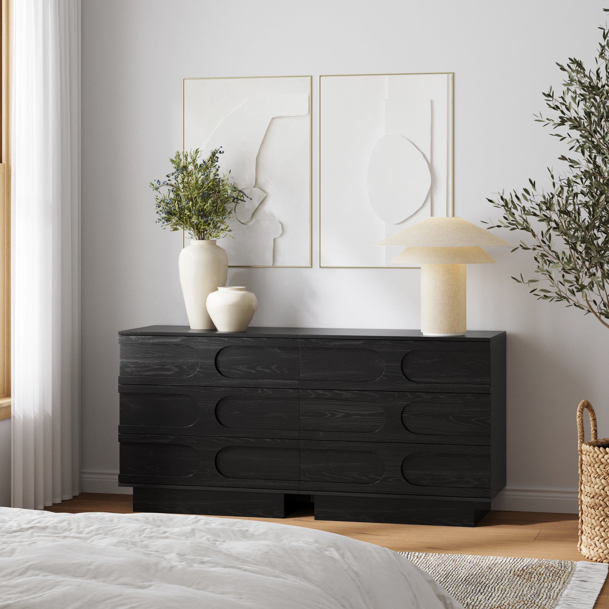 A modern bedroom showcases the Nathan James Mid-Century 3-Drawer Arched Dresser in black oak, accented with two green vases and a textured lamp. Above it hang two minimalist white art pieces, illuminated by natural light from the left.
