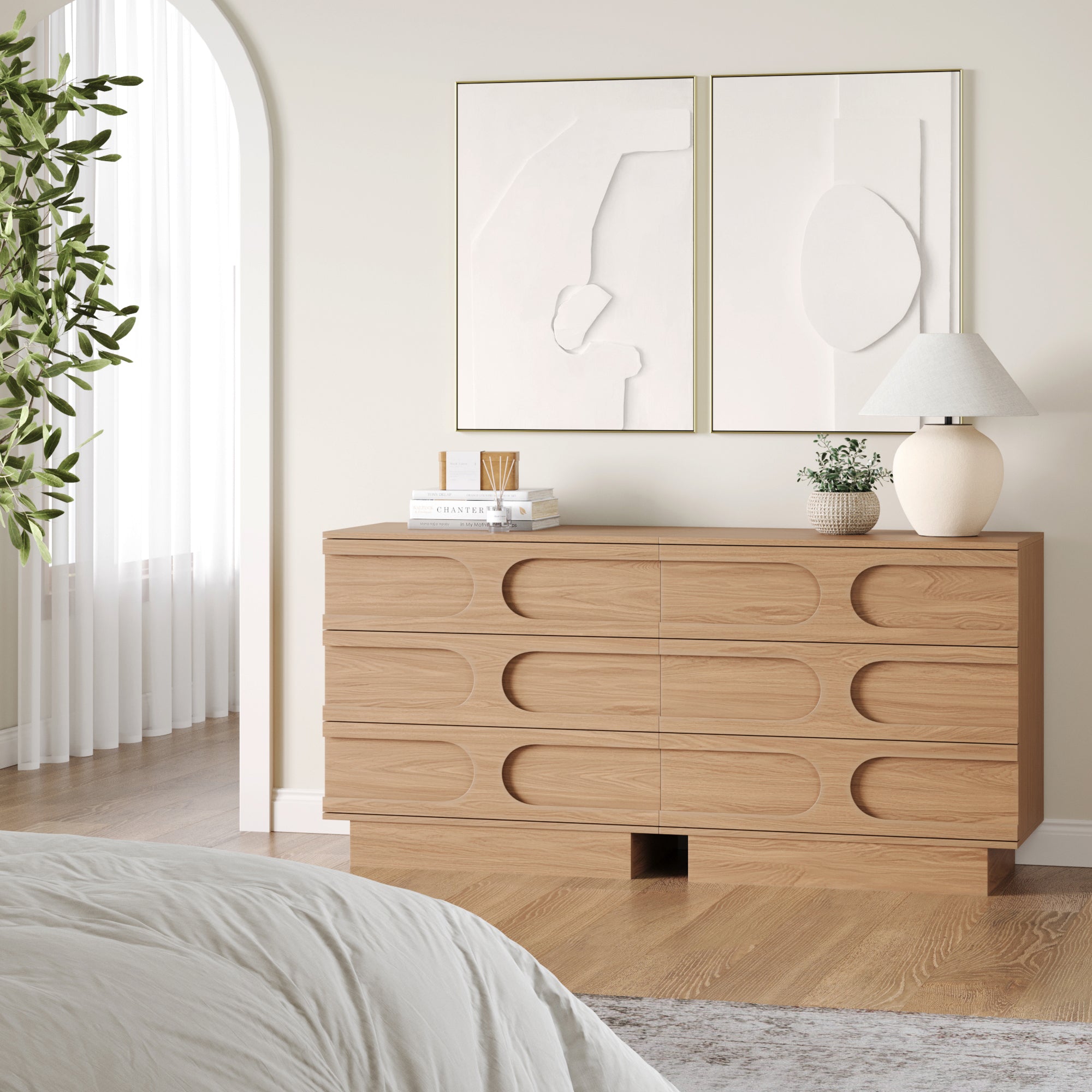 A modern bedroom showcases two Nathan James Mid-Century 3-Drawer Arched Dressers in raw oak, topped with a white lamp, books, and decor. Two abstract artworks hang above, and a leafy plant sits near an arched doorway with sheer curtains.