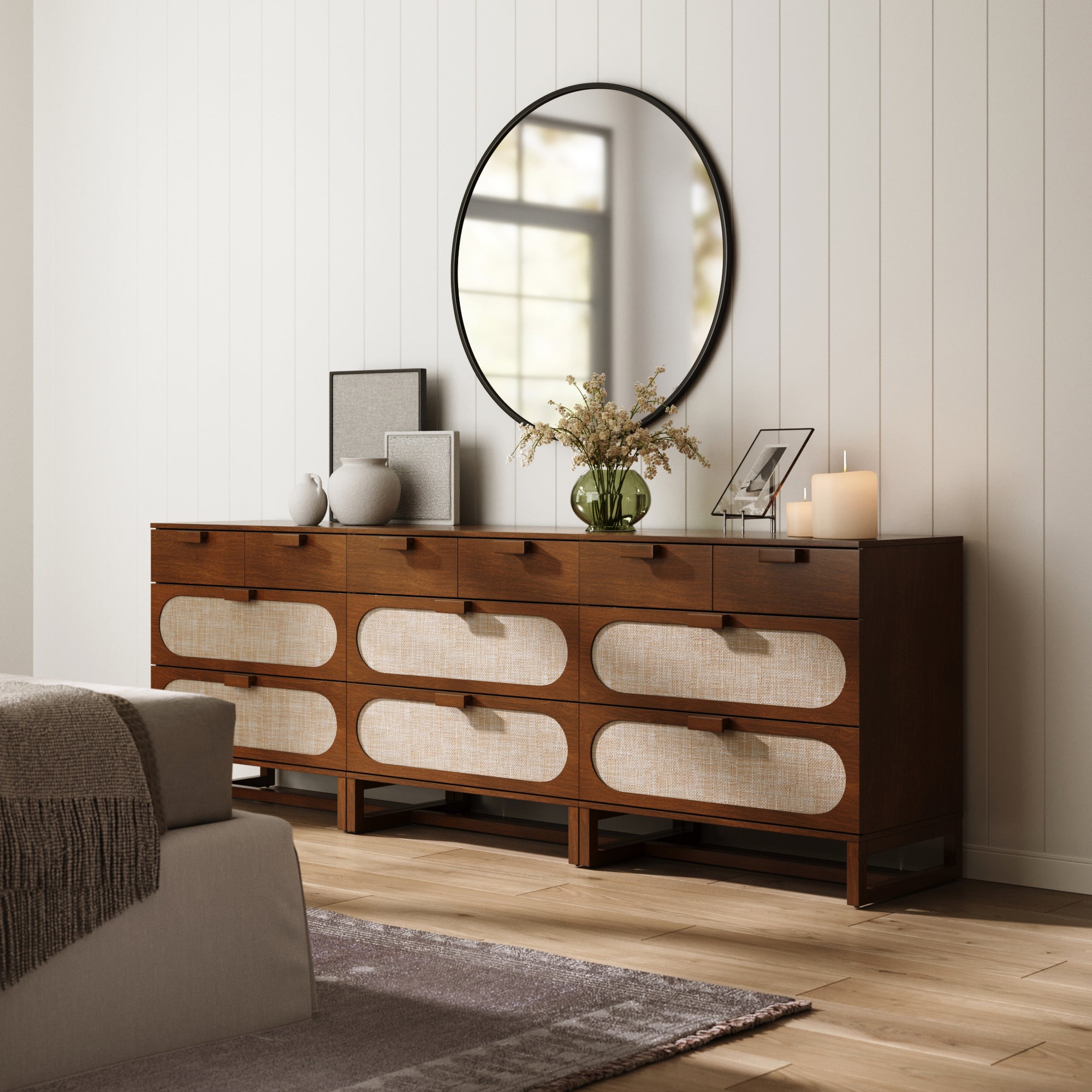 A set of three Nathan James Mid-Century Rattan 3-Drawer Dressers in Dark Acacia stands against a white paneled wall, topped with vases, framed photos, a candle, and flowers in a green glass vase beneath a large round mirror.