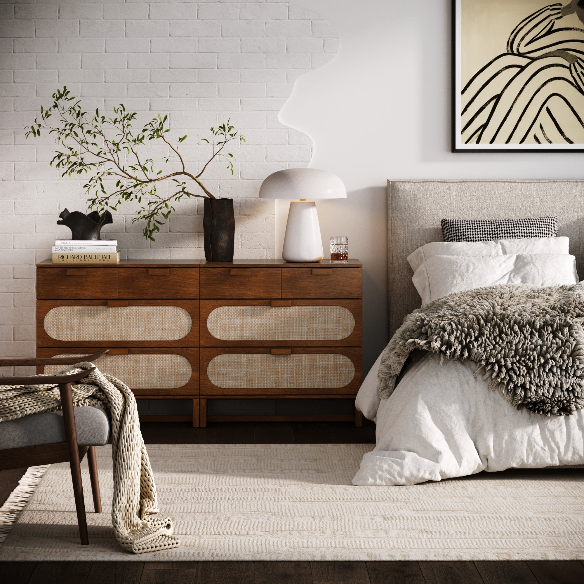 A cozy, modern bedroom with a Nathan James Mid-Century Rattan 3-Drawer Dresser in Dark Acacia, decorative accents, a bed with textured bedding, a gray chair with a knit blanket, and a potted branch against a white brick wall for mid-century flair.