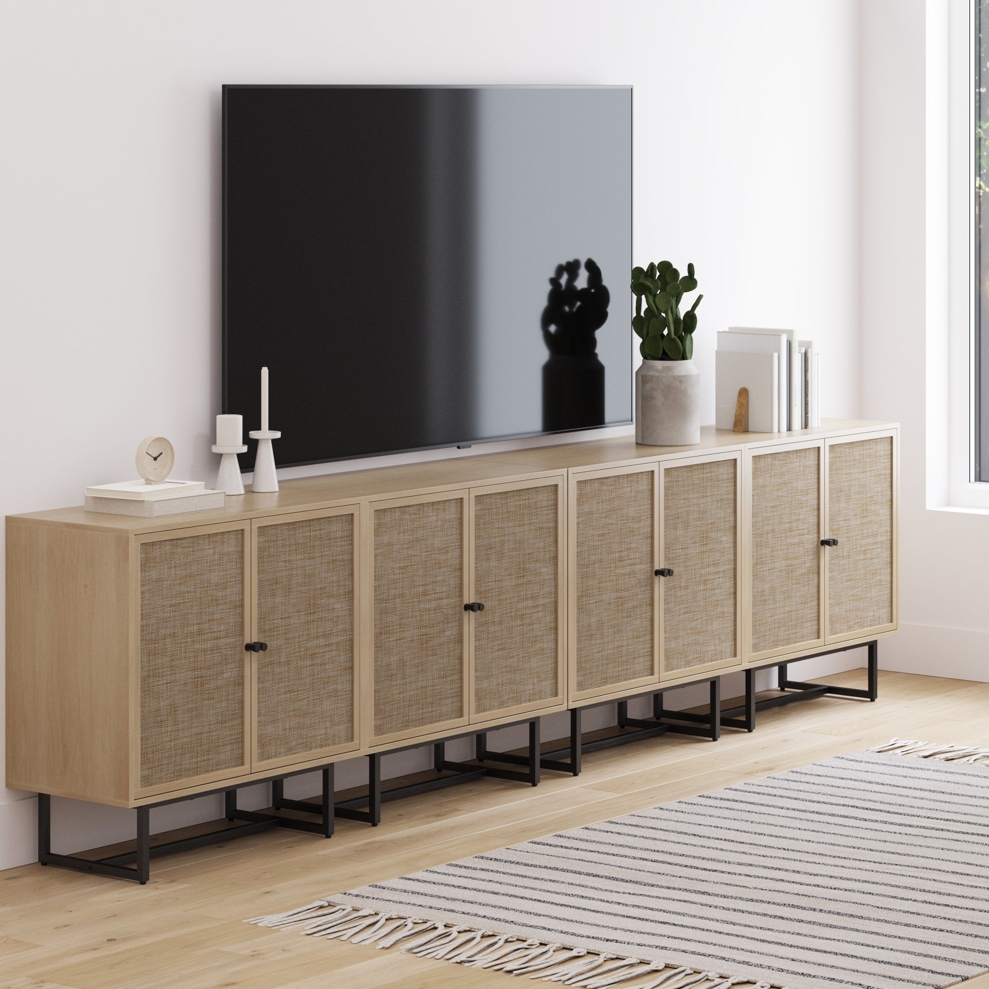 A modern living room showcases a flat-screen TV on the Nathan James Large Rattan Credenza Console Table Oak (Set of 4), styled with candles, a clock, books, and a plant. A striped rug complements the cozy, bohemian atmosphere.