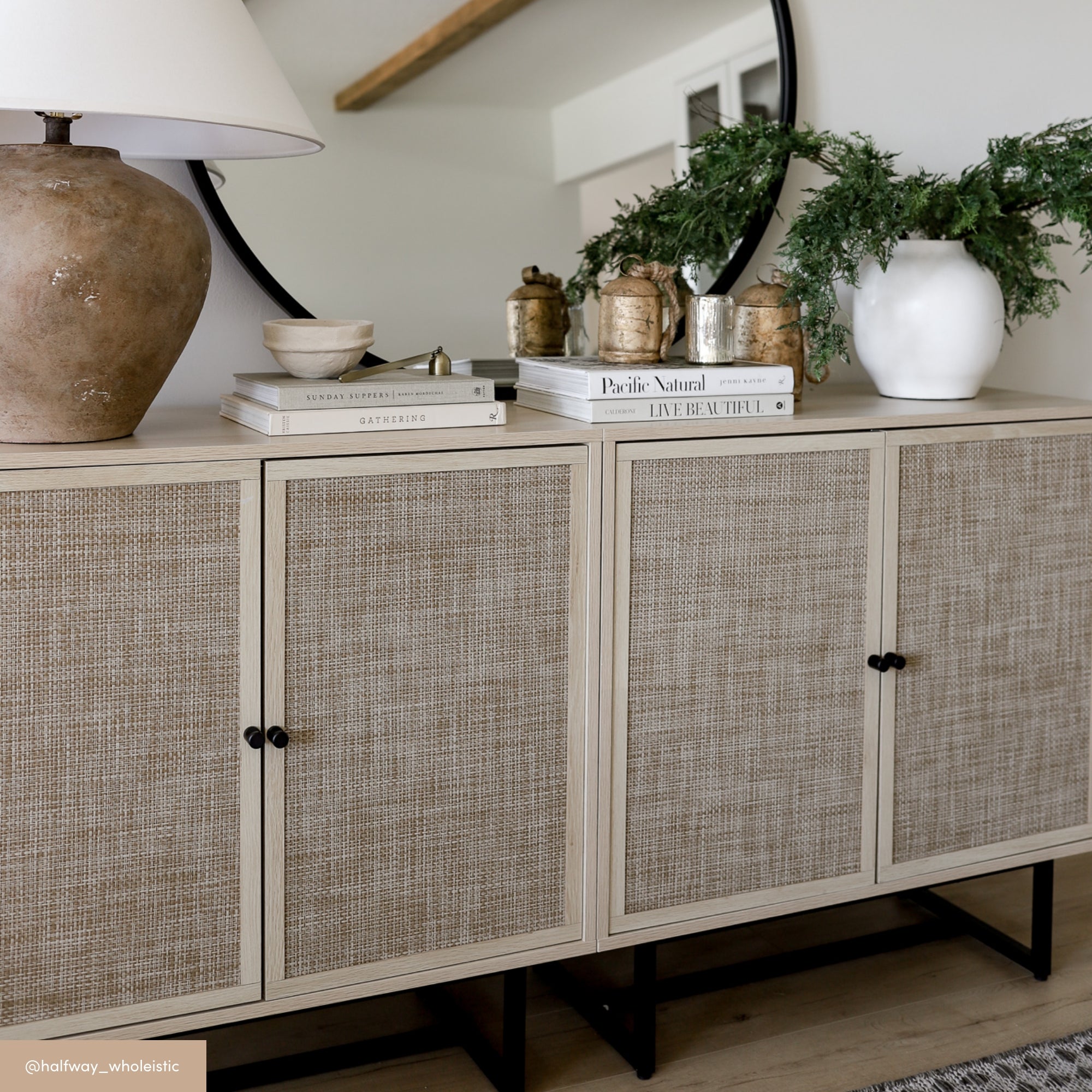 A light wood sideboard with woven doors, topped with a ceramic lamp, stacked books, glass jars, and a white vase holding greenery, sits in front of a round mirror on a light-colored wall.