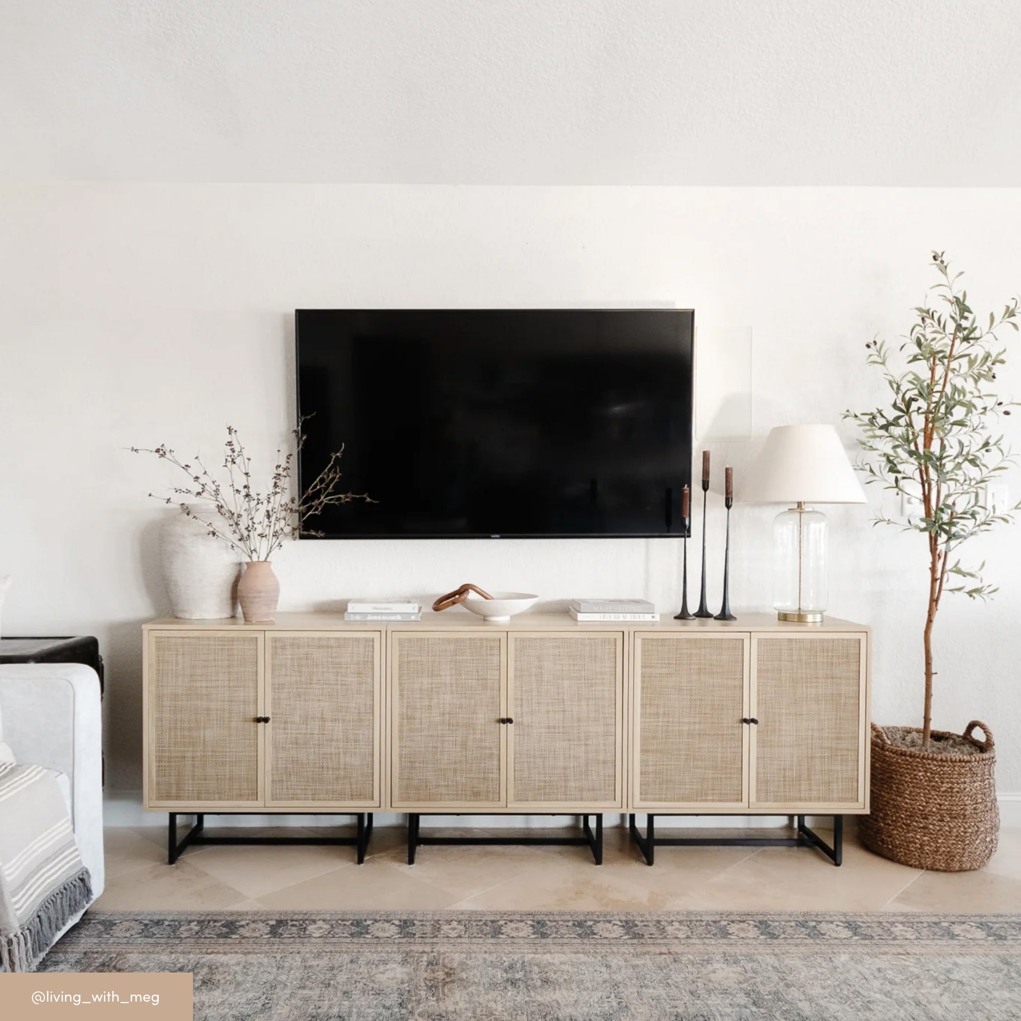 A minimalist living room features a rattan-front sideboard with decor items, including vases, books, candles, and a lamp, beneath a wall-mounted flat-screen TV. A tall potted plant stands to the right on a neutral rug.