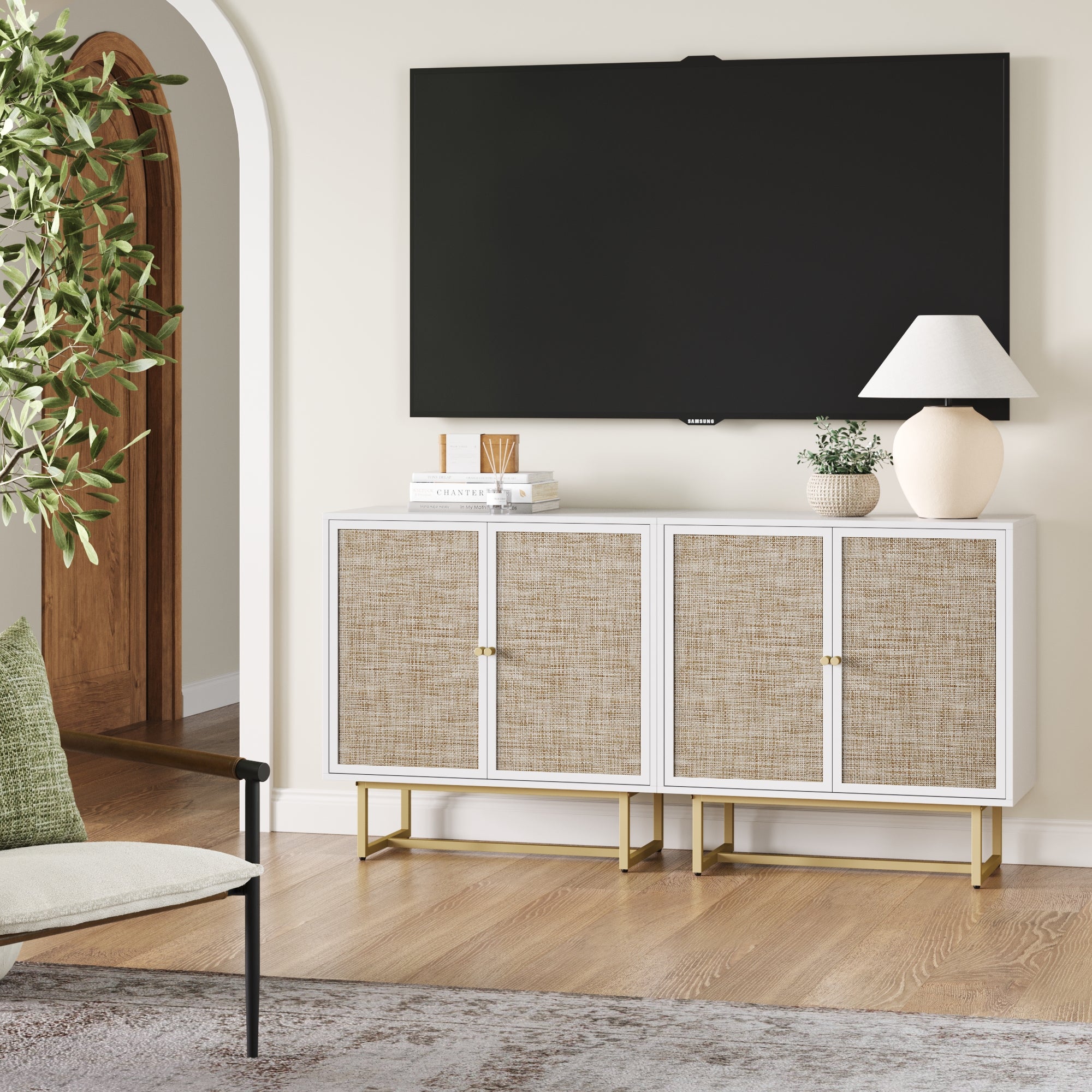 A modern living room showcases a wall-mounted TV above the Nathan James Rattan TV Console Credenza White (Set of 2) with white and rattan doors and gold legs, topped with a lamp, vase, and books. A potted plant and chair sit to the left.