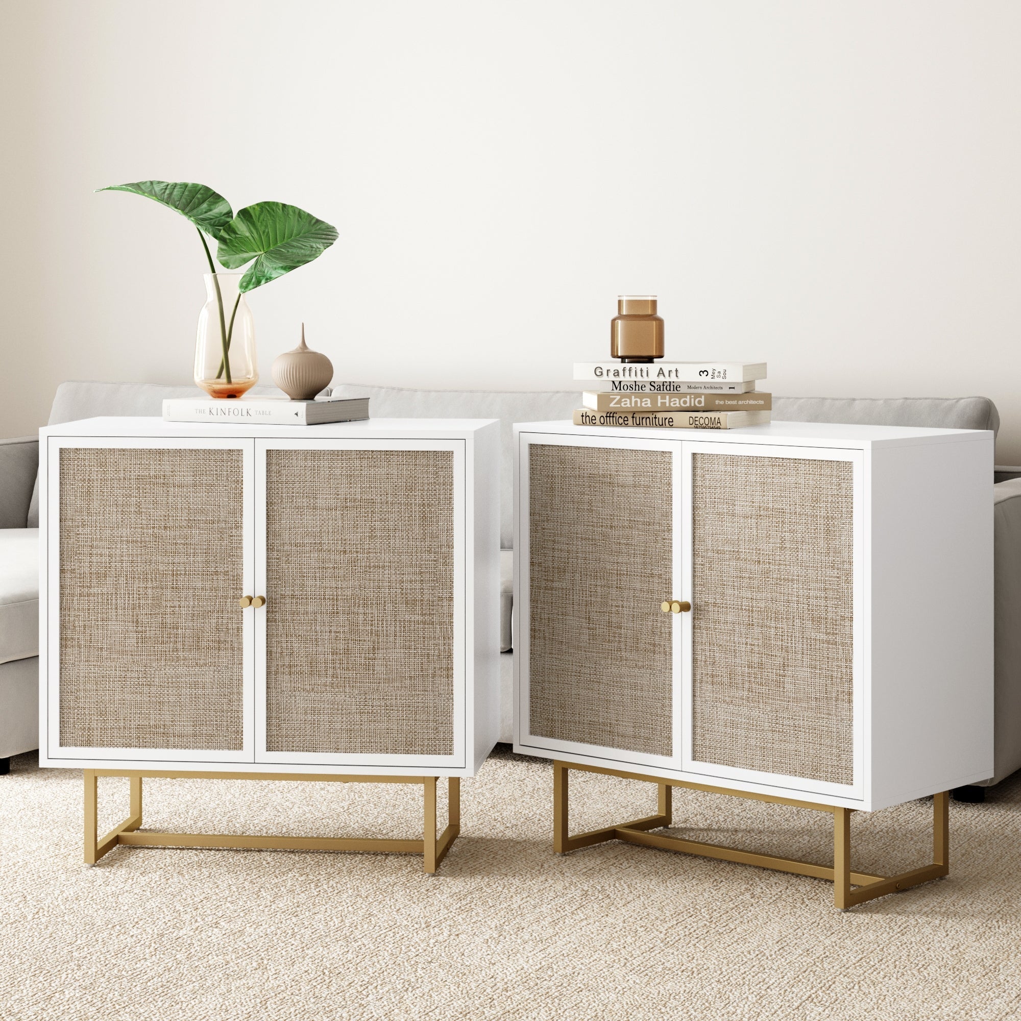 Two white cabinets with woven rattan doors and gold legs sit side by side in a living room. Decorative vases, a green leaf, and stacked books are placed on top. A neutral sofa and cream carpet are in the background.