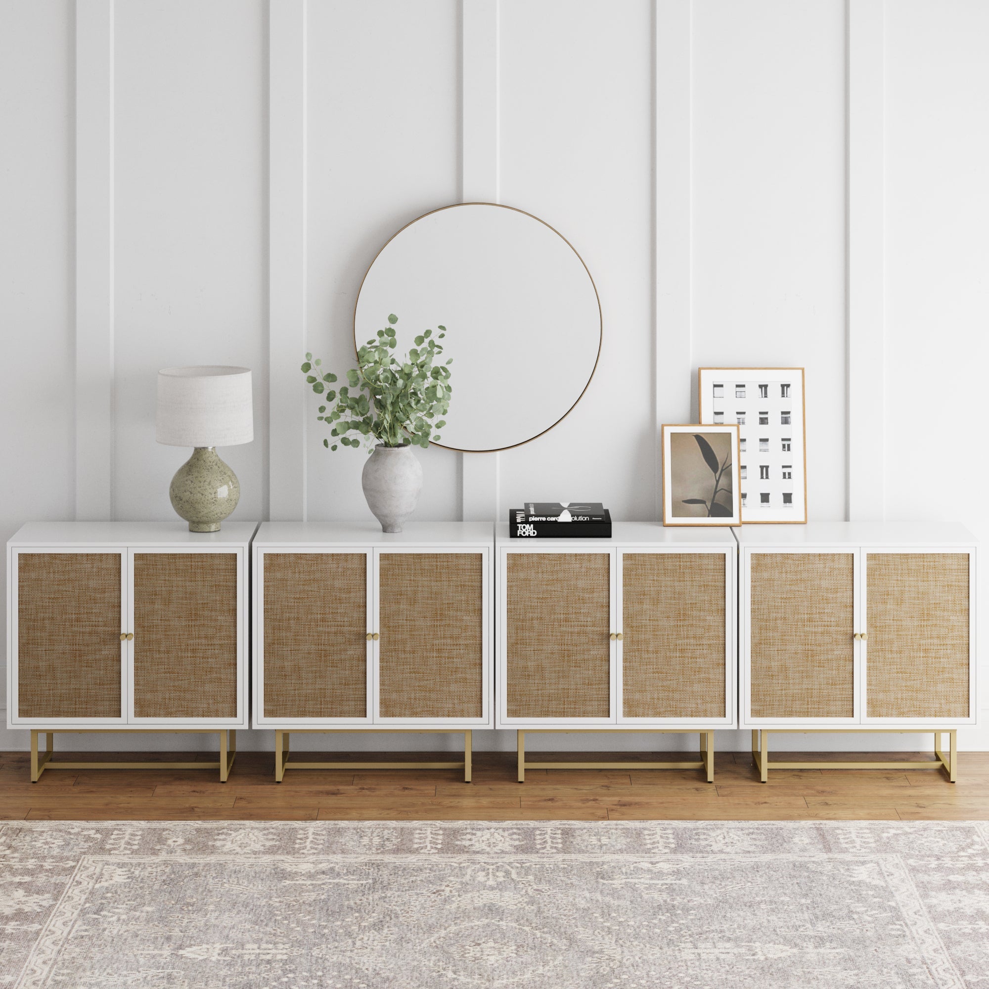 The Nathan James Large Rattan Credenza Console Table (Set of 4) sits against a white paneled wall and patterned rug, topped with a round mirror, ceramic lamp, greenery-filled vase, books, and framed art on a wooden floor.