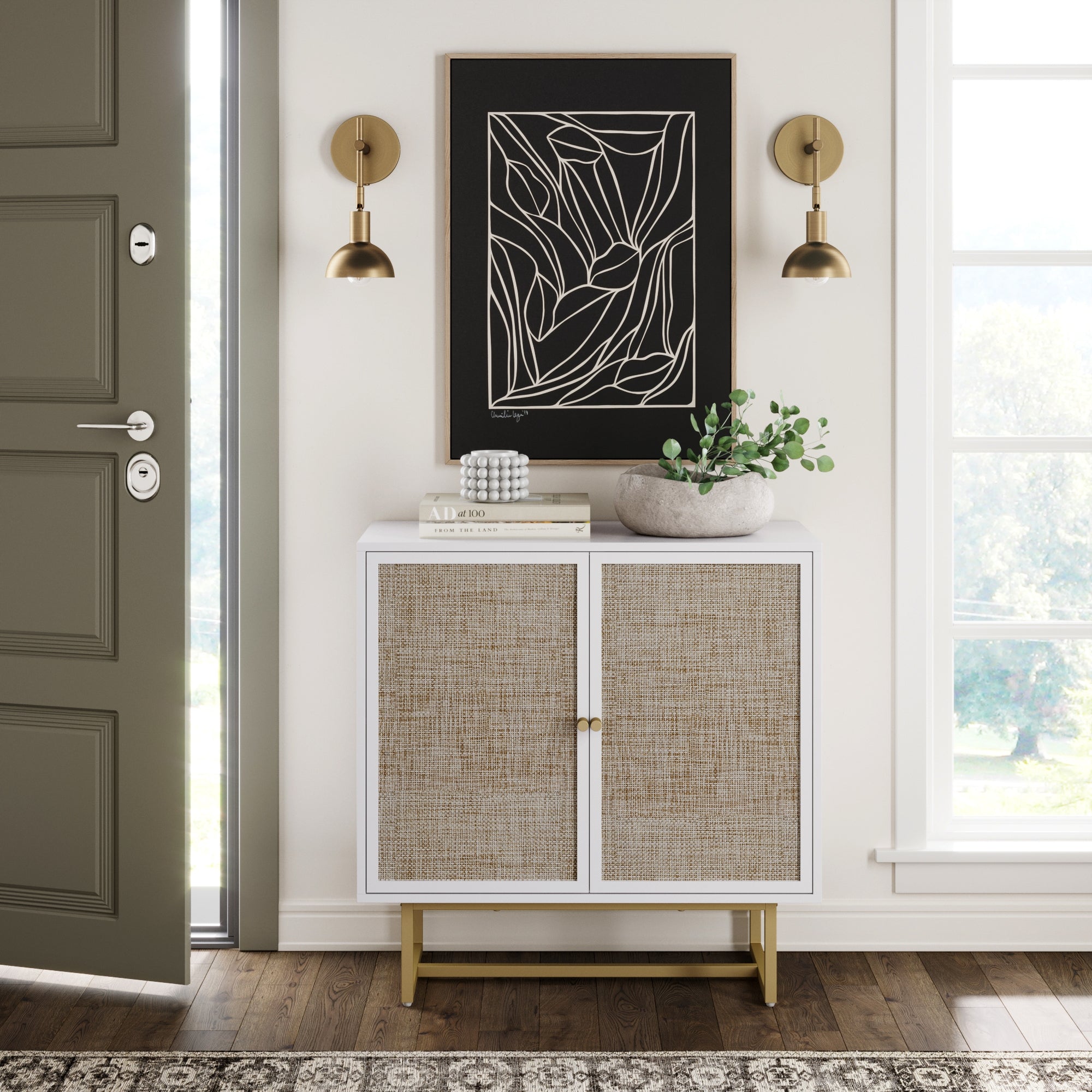 A modern entryway with an olive green door, potted plant, decor books, abstract wall art, brass sconces, a large window, and the Nathan James Wood & Rattan Storage Cabinet in white featuring woven doors and an adjustable shelf.