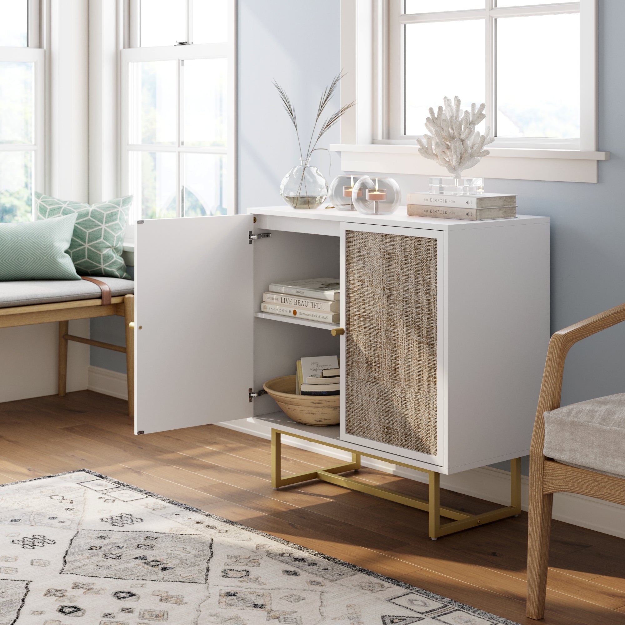 A modern white cabinet with one open door, featuring woven rattan detail, gold legs, and shelves with books and decor. It sits in a bright room with large windows, a bench, and light wood flooring.