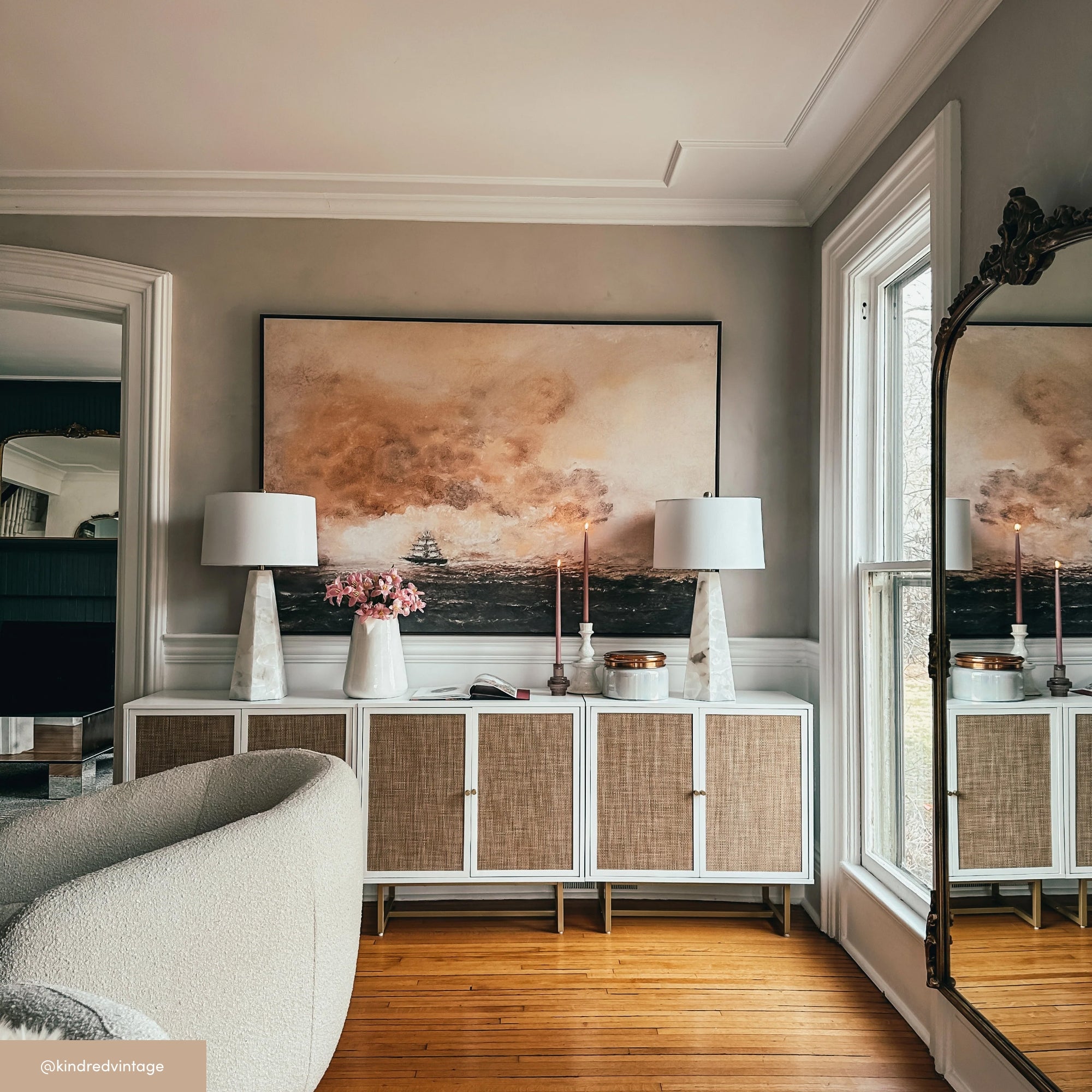 A stylish living room features a white sideboard with wicker doors, topped with two white lamps, pink flowers, and candles. Above hangs a seascape painting, and a large mirror sits near tall windows.